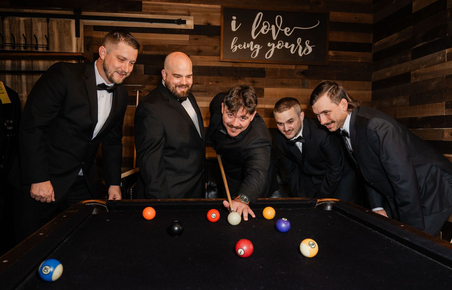 Five men in formal suits smiling around a pool table with colorful billiard balls at a wedding venue.