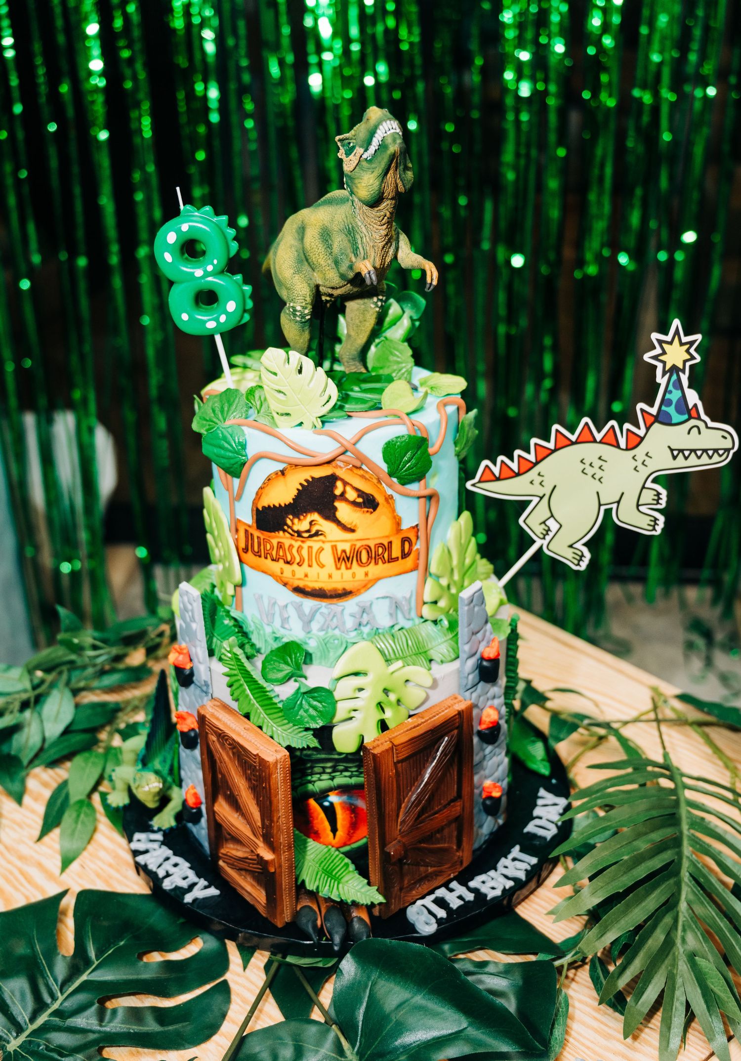 Jurassic World themed 2-tier birthday cake with T-Rex topper, tropical leaves, and number 8 candle for an 8th birthday.