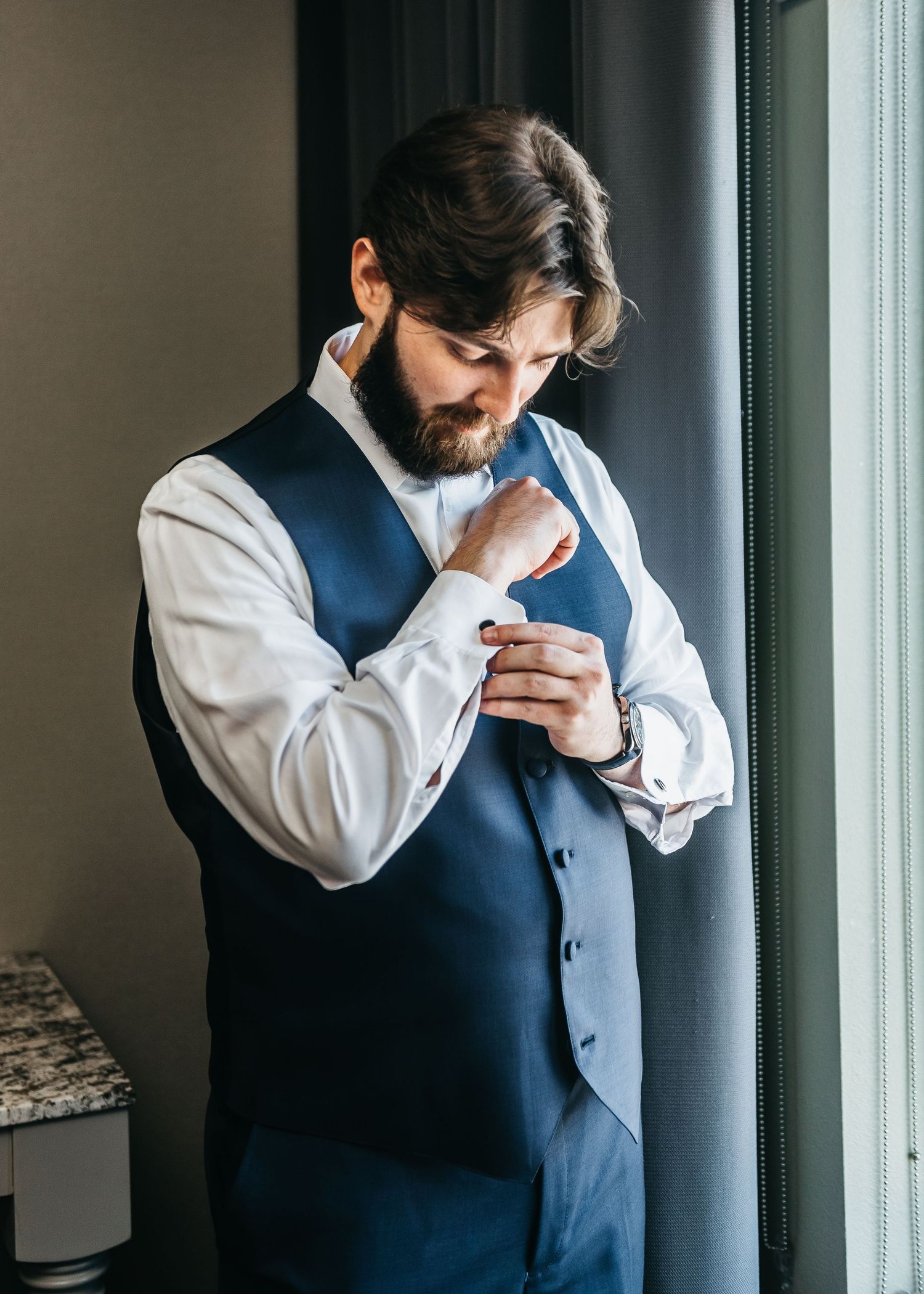 A well-dressed groom in a navy vest and white shirt adjusts his cufflinks near a window.