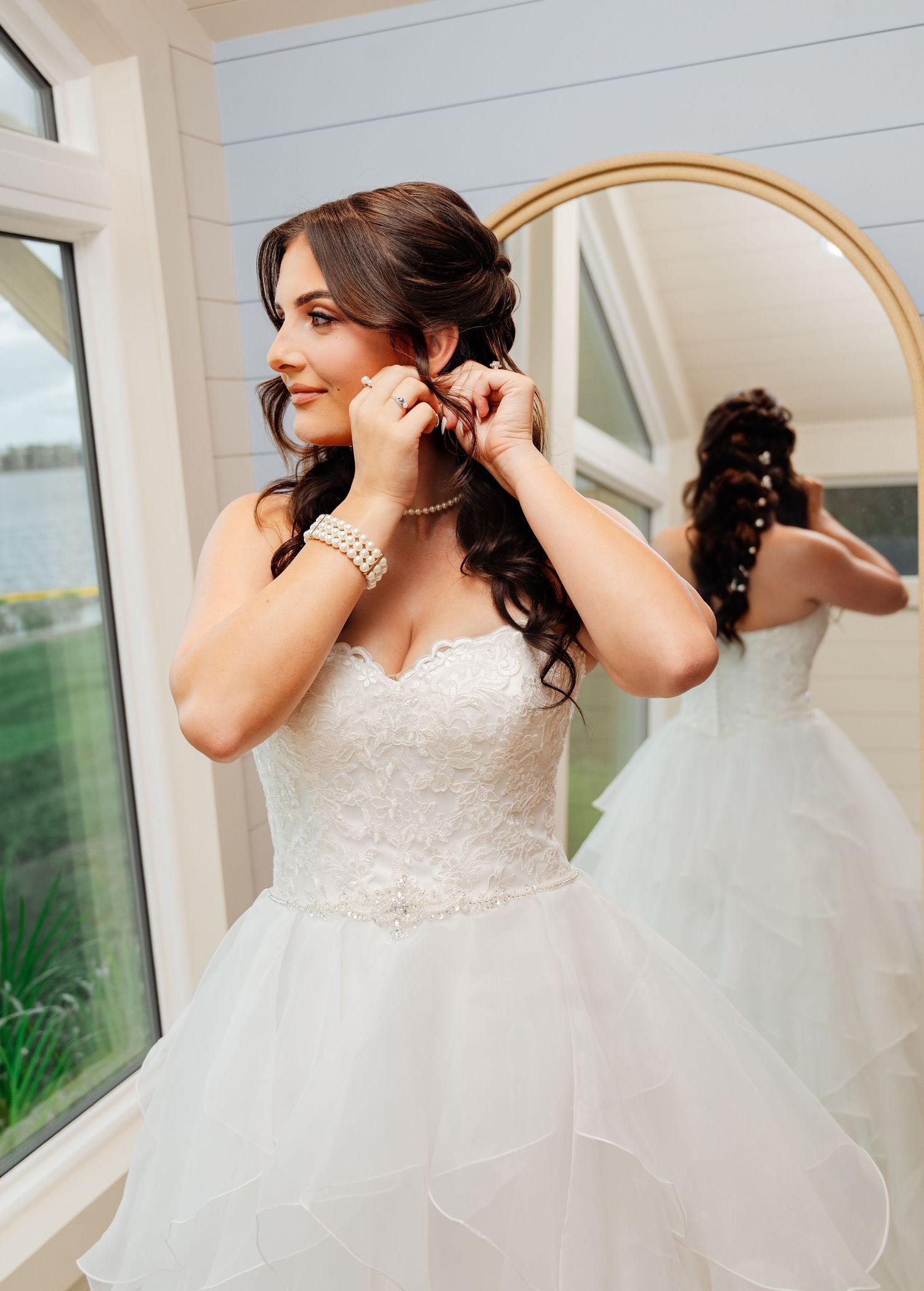 Bride in strapless lace ballgown adjusting pearl earrings near mirror, wearing pearl bracelet and necklace.