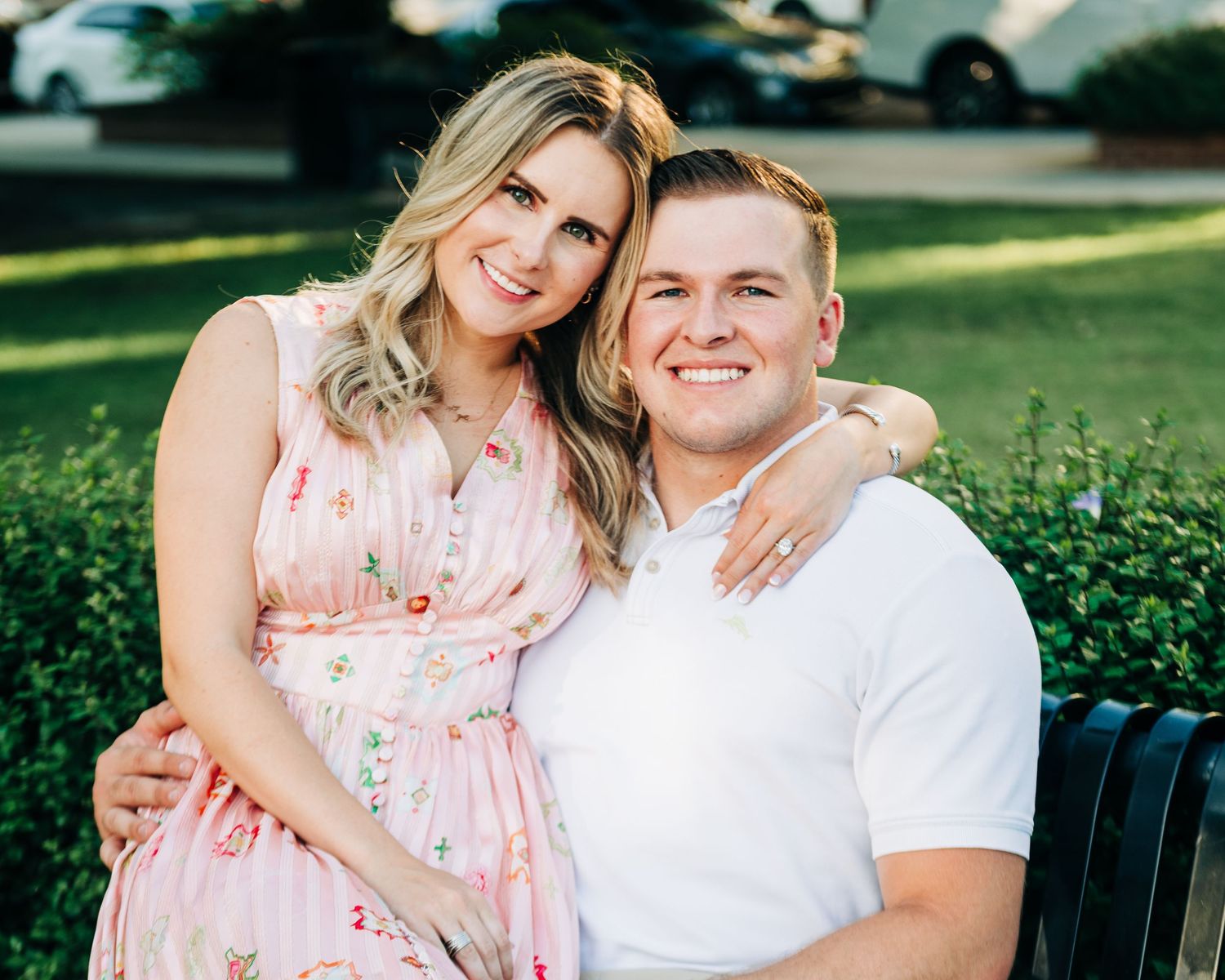 Happy couple smiling together outdoors, woman in floral dress embracing man in white polo on a park bench.