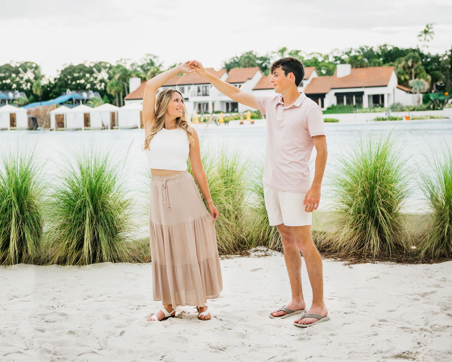 Couple dancing on a sandy beach near a waterfront property surrounded by tropical greenery.