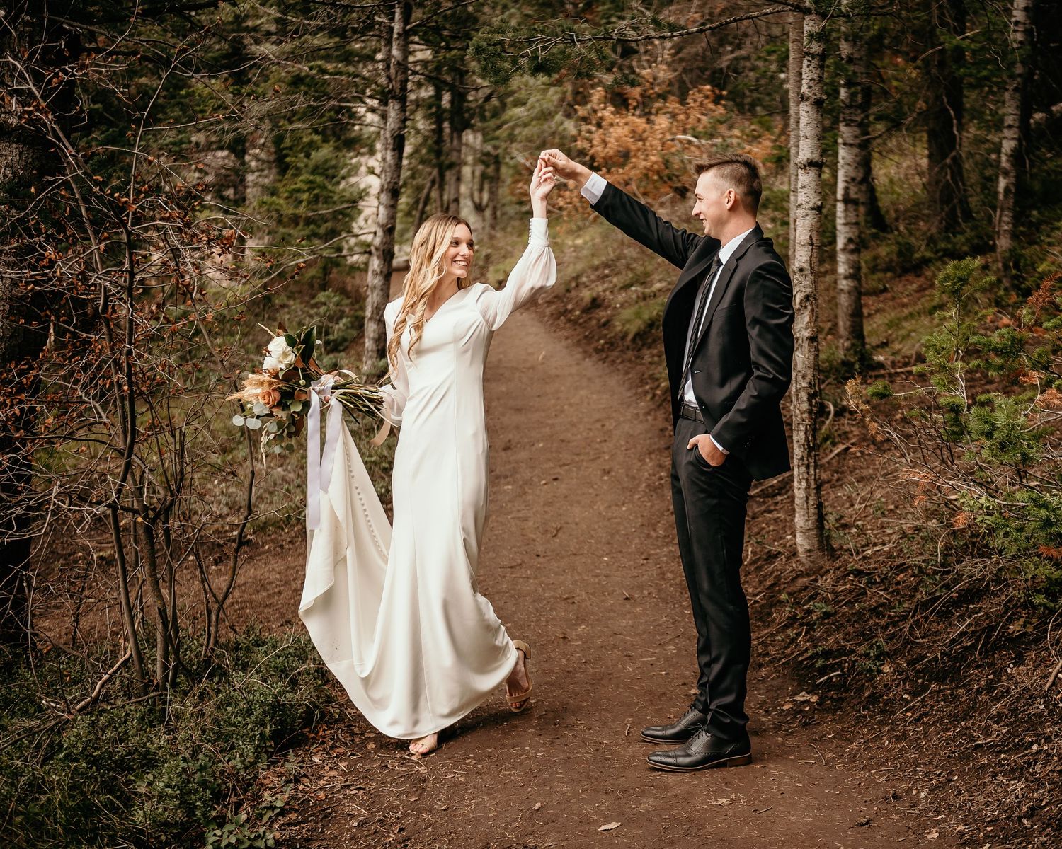 Bride and groom dancing on a forest path, bride in white gown holding bouquet, groom in black suit.