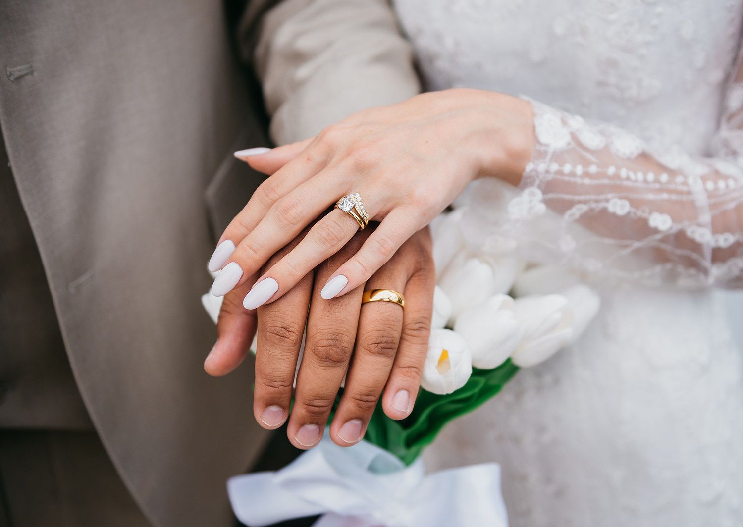 Newlywed couple displaying wedding rings, bride holding white tulip bouquet in elegant lace dress.