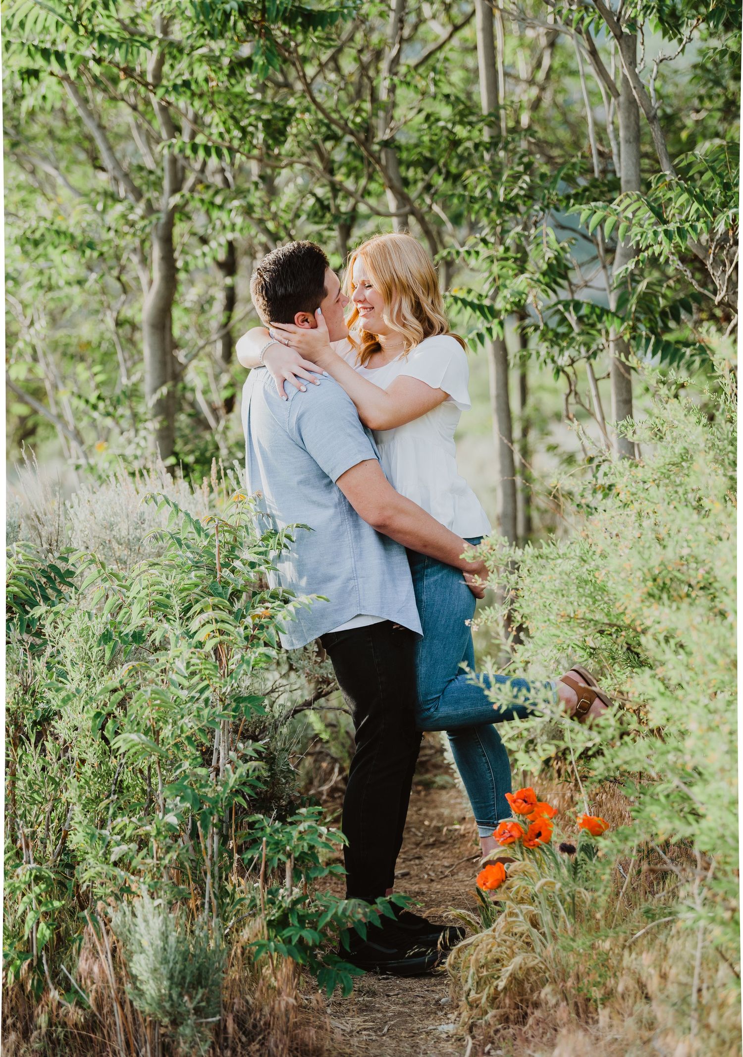 Couple embracing and kissing on a forest trail surrounded by lush greenery and orange wildflowers.