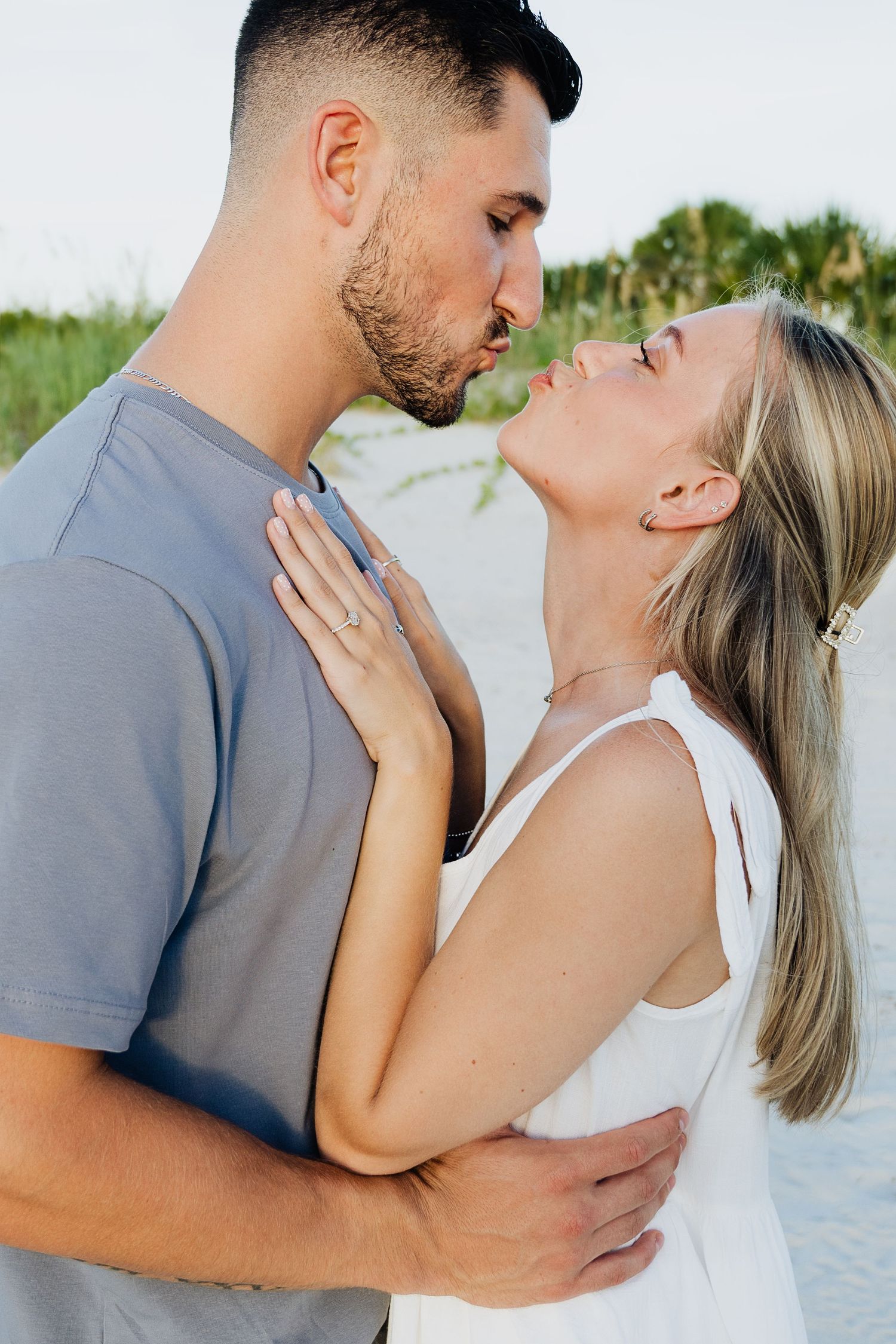 Couple sharing a romantic kiss on a sandy beach, woman wearing white dress and engagement ring.