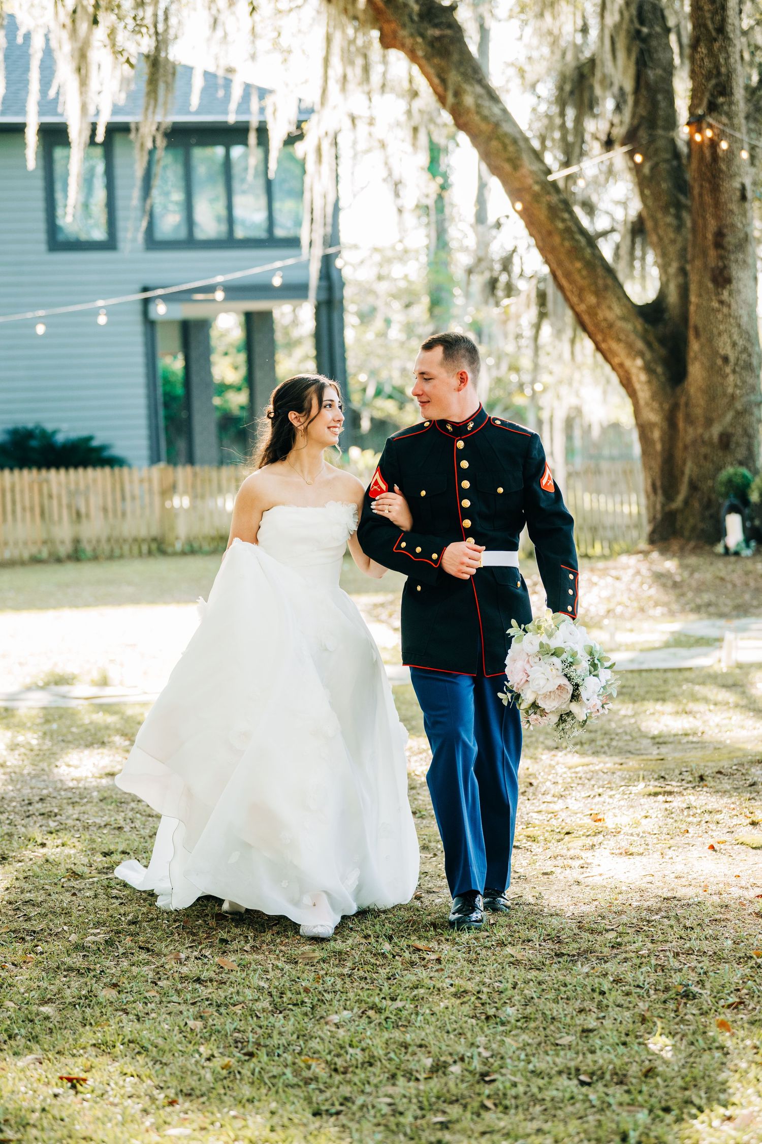 A bride in a white gown and groom in Marine dress blues walk together outdoors under Spanish moss-draped oak trees.
