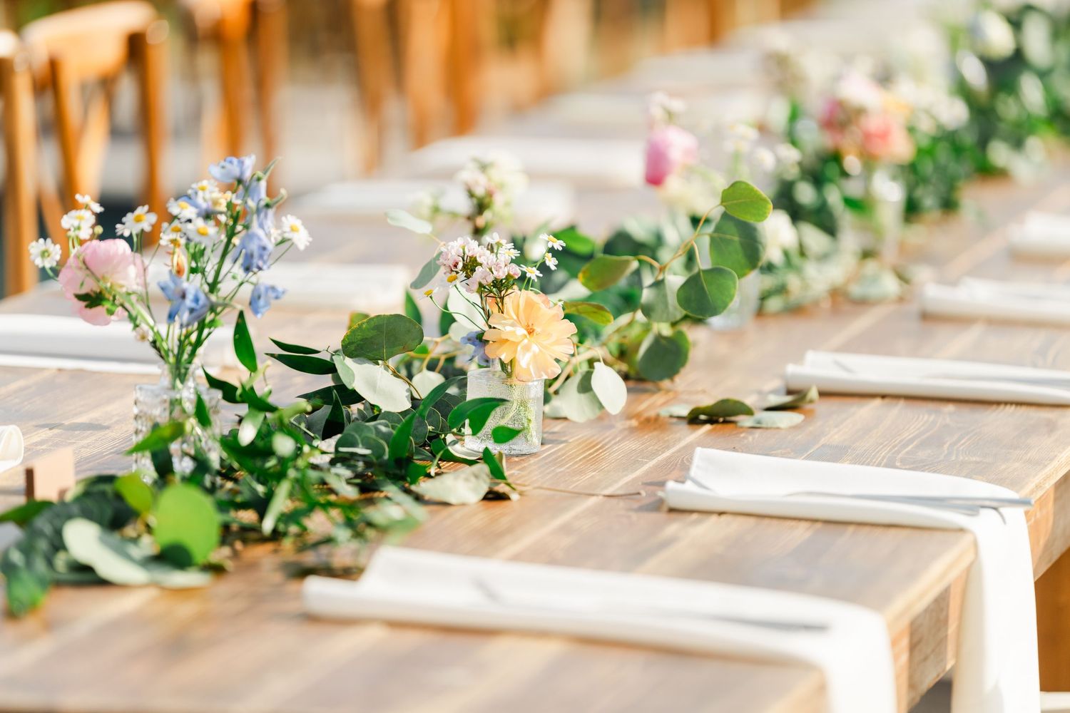 Rustic wooden wedding reception table with greenery garland and colorful wildflowers, white napkins, and elegant place settings.