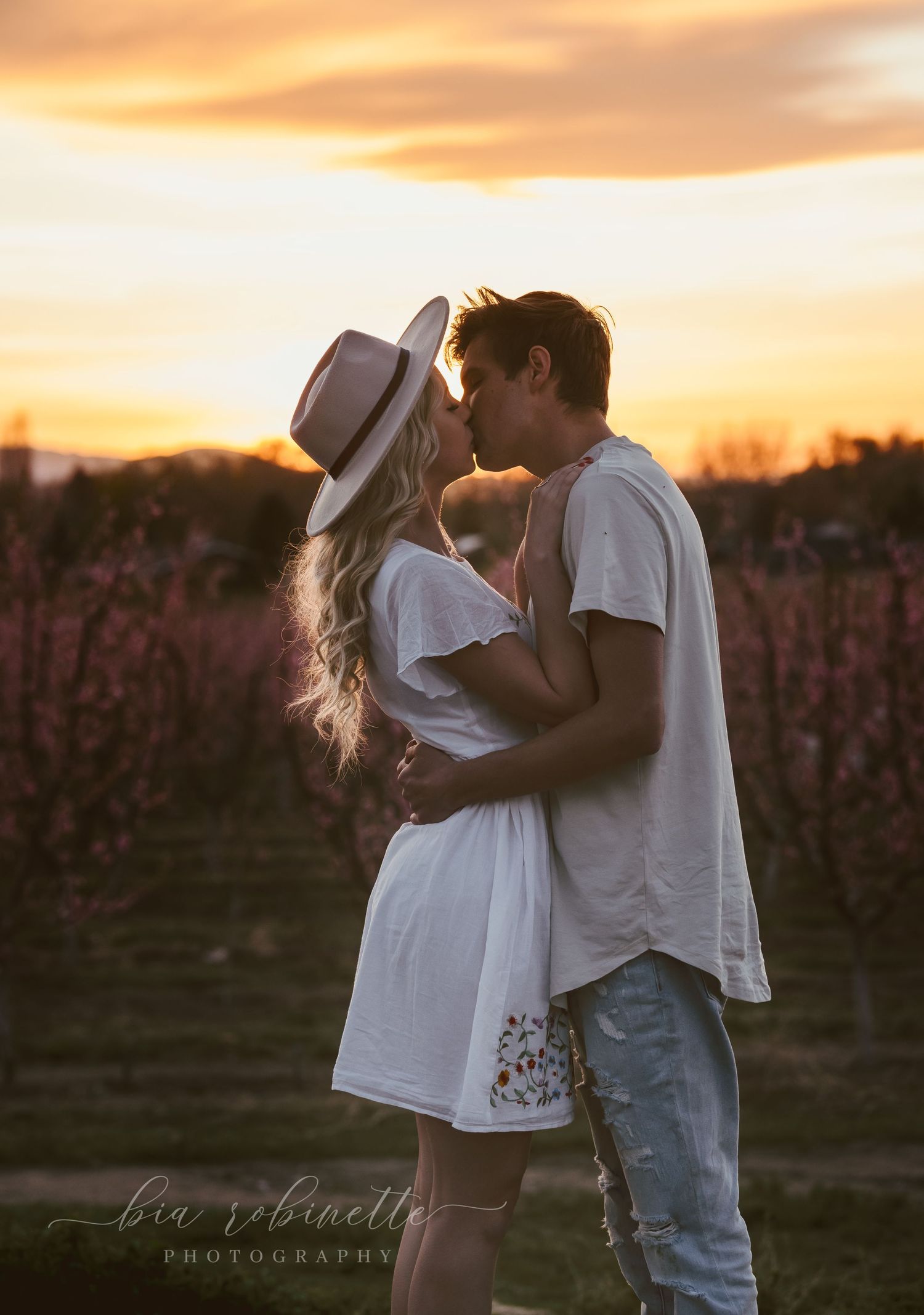 Couple sharing a romantic kiss at sunset in a vineyard, woman in white dress and hat, golden sky backdrop.