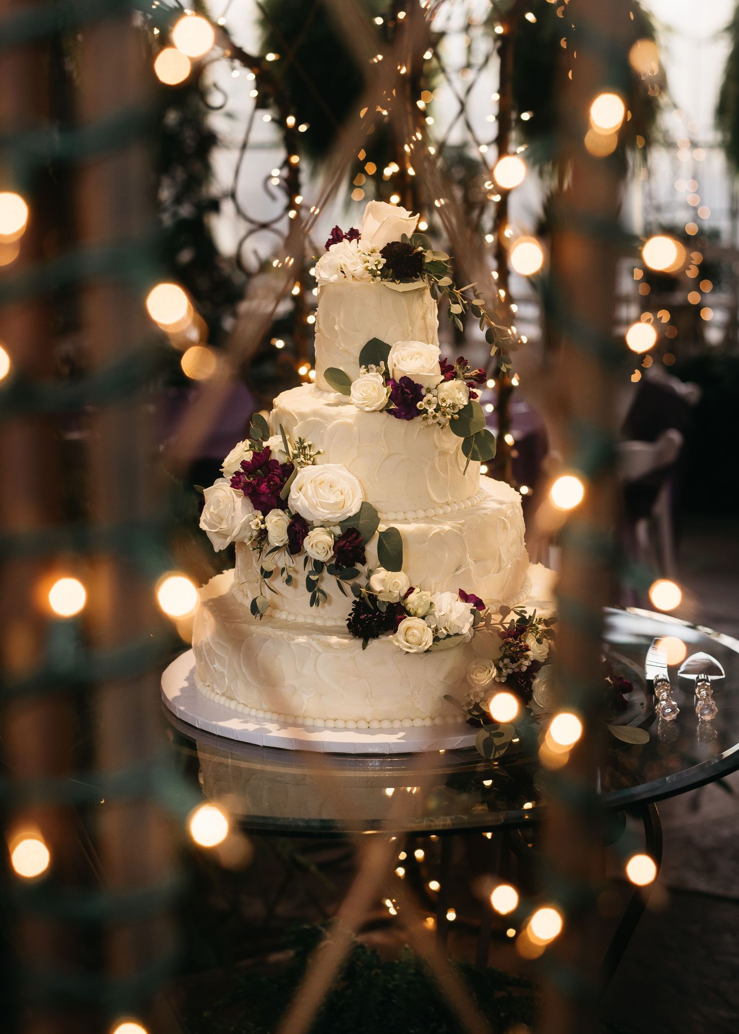 Three-tier white wedding cake adorned with burgundy and ivory roses, surrounded by warm bokeh string lights.