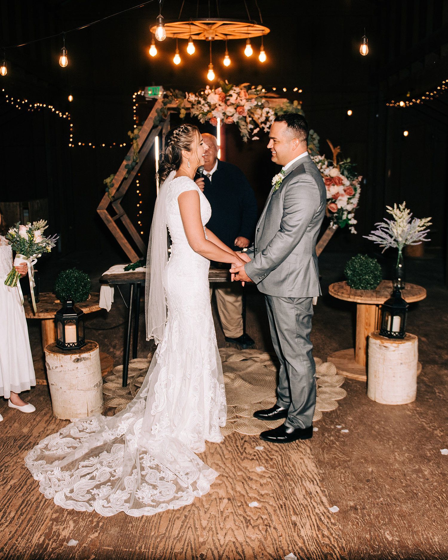 Bride and groom hold hands at rustic outdoor wedding ceremony with string lights and floral arch backdrop at night.