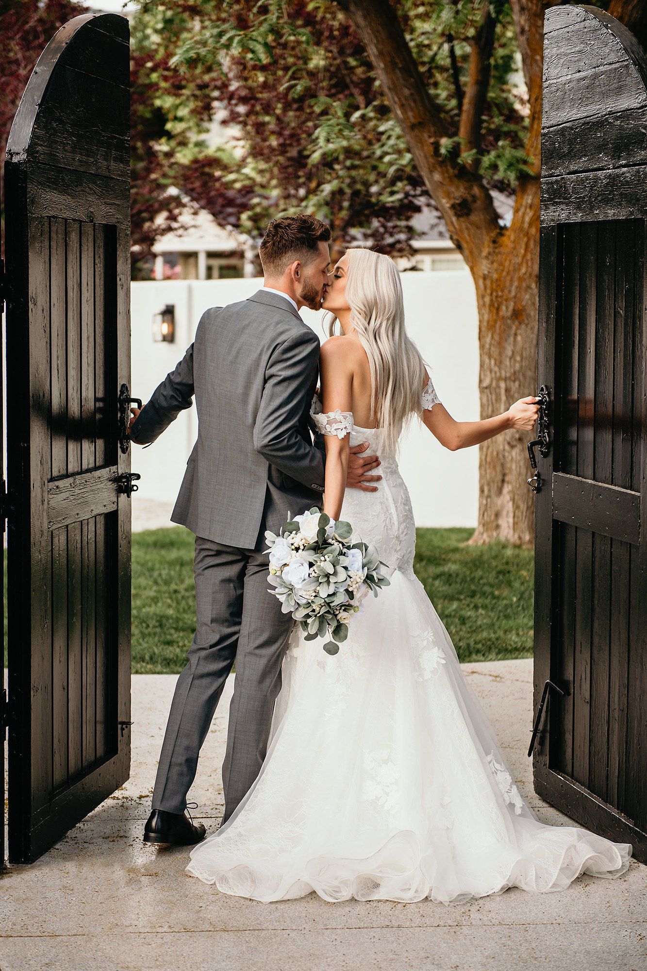 A bride and groom share a kiss while opening elegant black wooden gates, she holds a white floral bouquet.