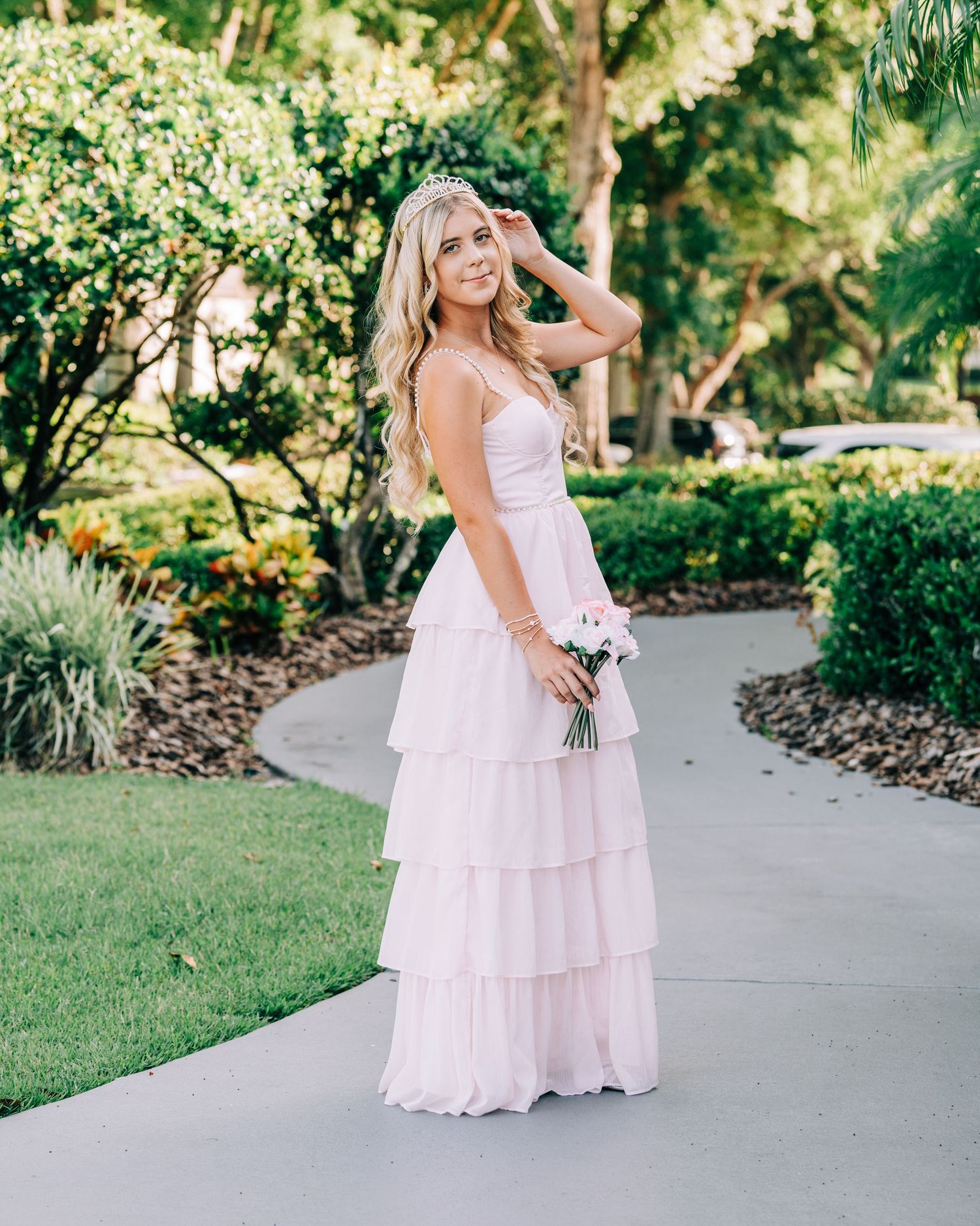 Blonde woman in a blush pink tiered maxi dress holding flowers, posing on a garden pathway surrounded by lush greenery.