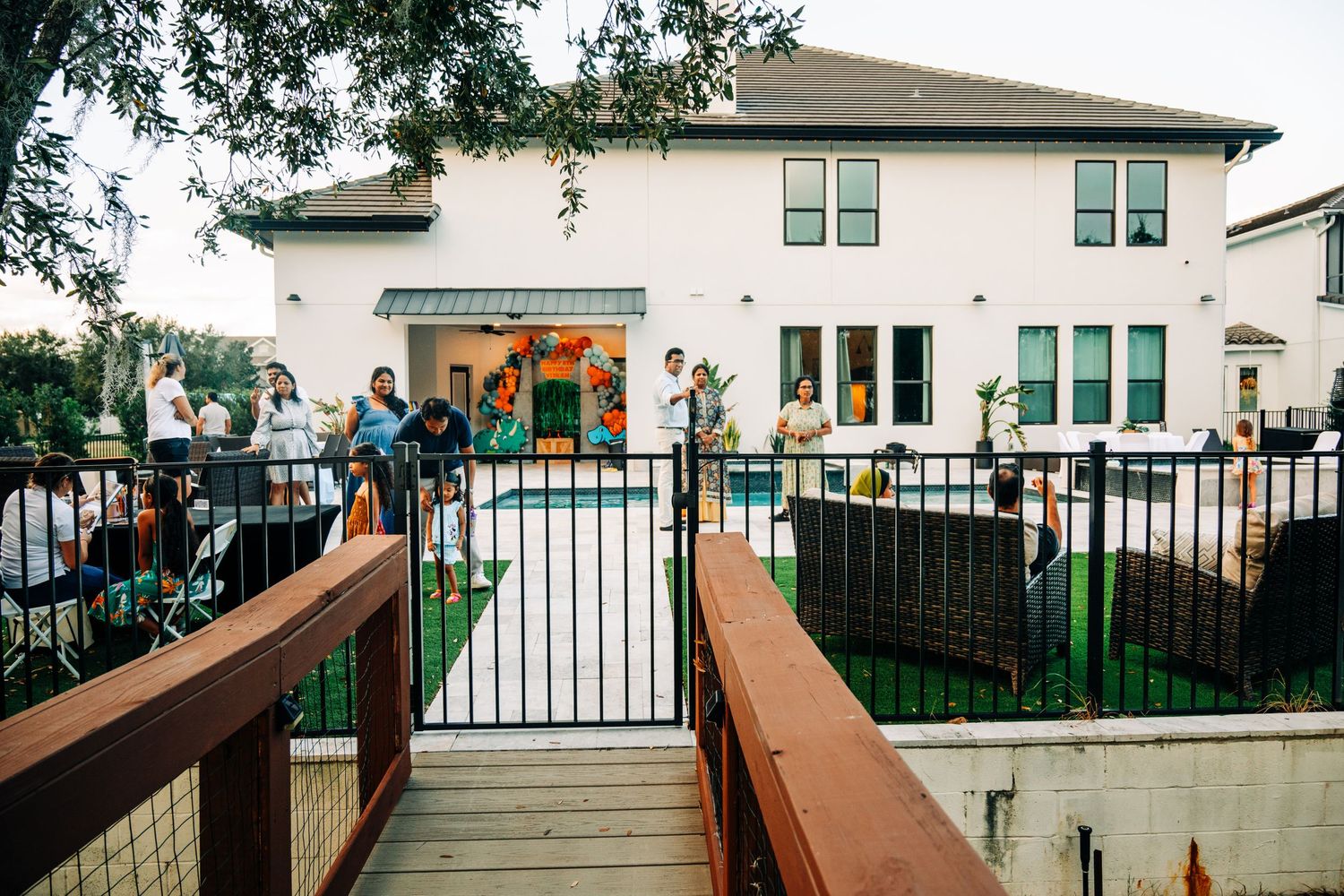 Guests gather around a fenced backyard pool at a house party, with a white two-story home and decorations visible.