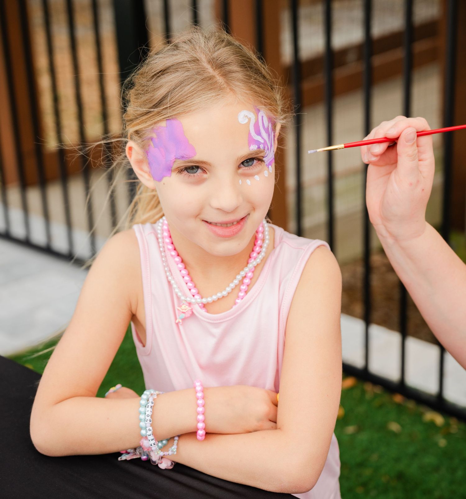 Young girl smiling while getting purple butterfly face paint applied at a party, wearing pink dress and pearl jewelry.