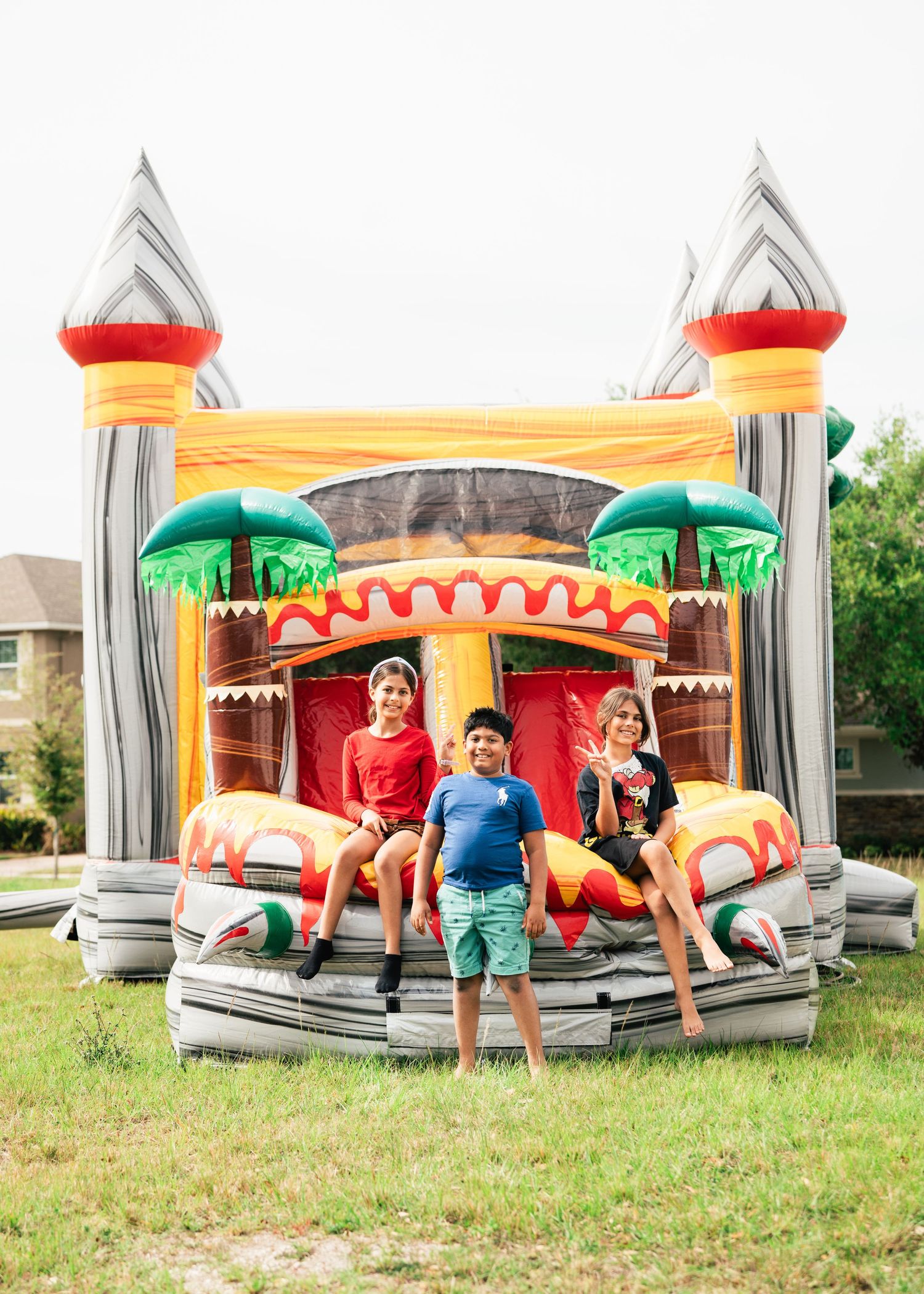 Three children sitting on a colorful inflatable bounce house castle in a grassy backyard.