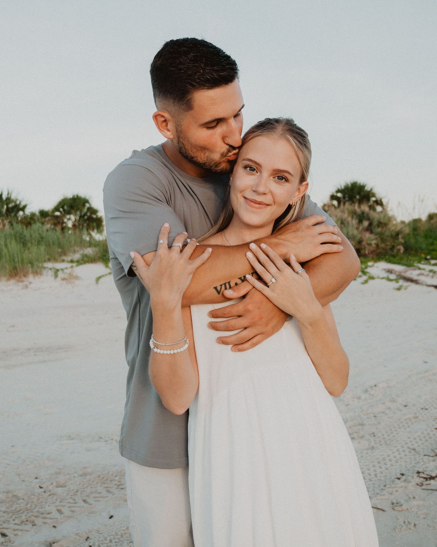 Couple embracing on a sandy beach, man kissing woman's head, she wears a white dress at sunset.