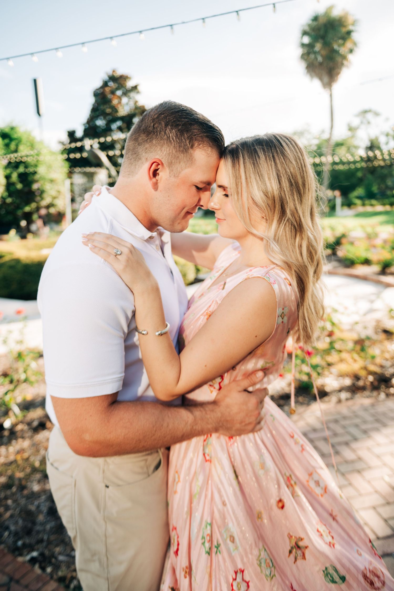 Couple sharing a romantic embrace outdoors, woman in floral pink dress, string lights and greenery in background.