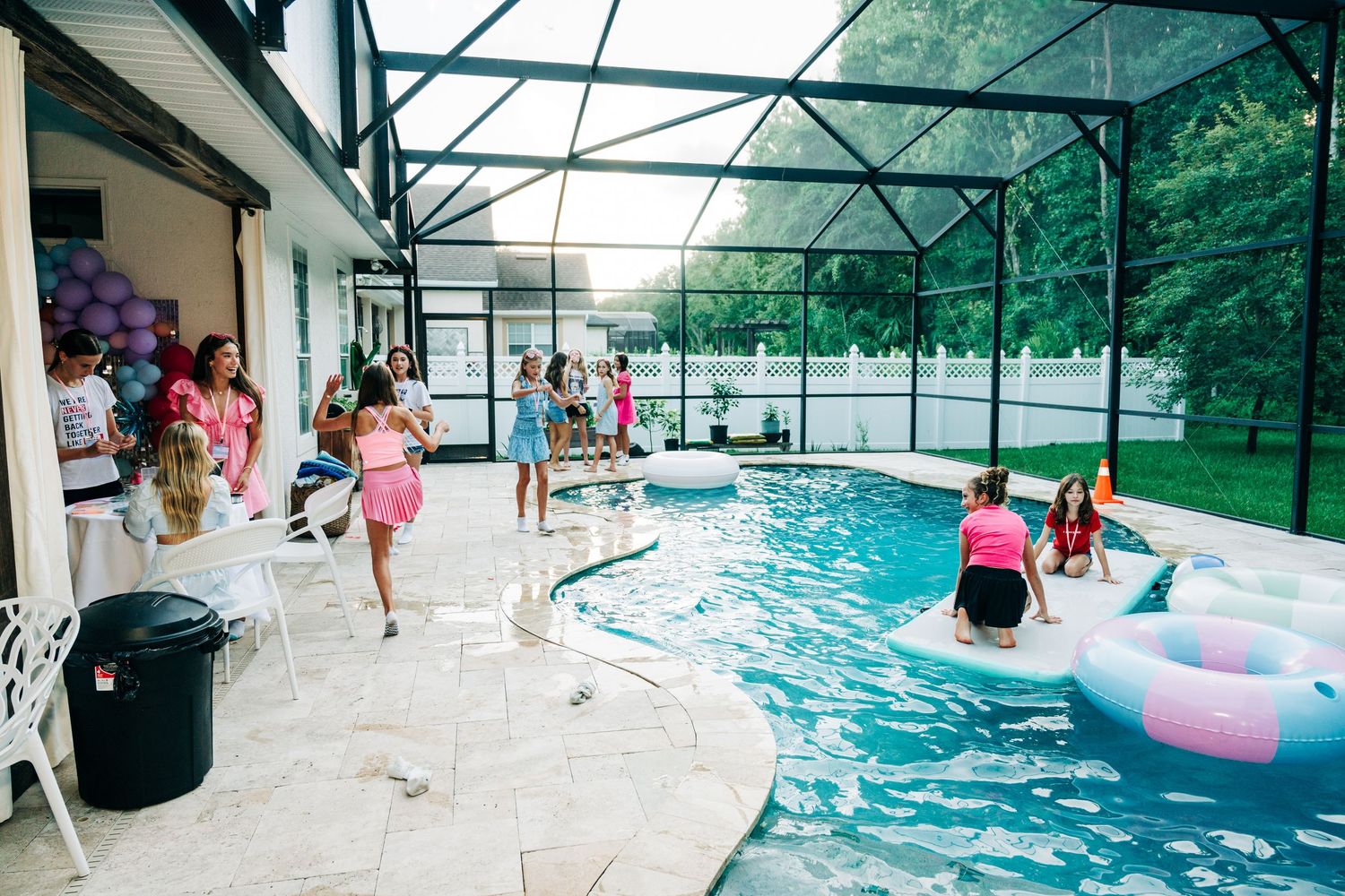 Girls enjoying a pool party under a screened enclosure, with floaties and purple balloons decorating the festive backyard.