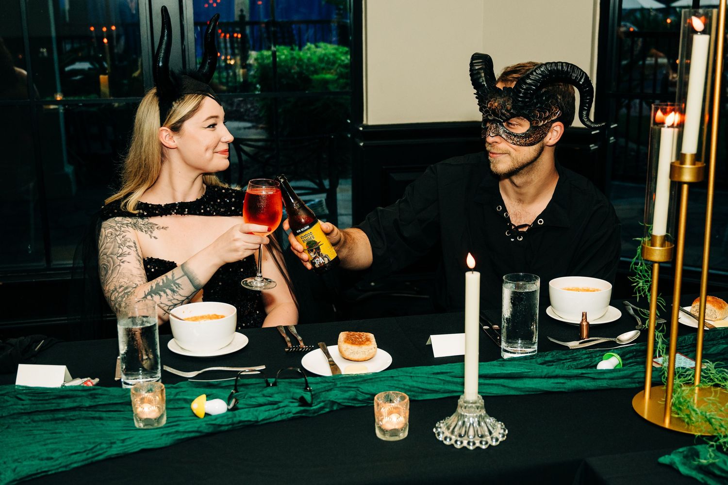 Couple in Halloween masks toasting with wine at a candlelit dinner table with green tablecloth.