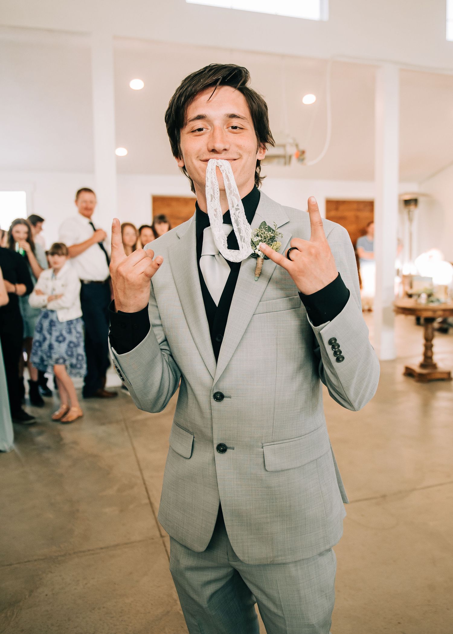 Smiling groom in gray suit holding wedding rings, flashing rock on signs at indoor wedding reception.