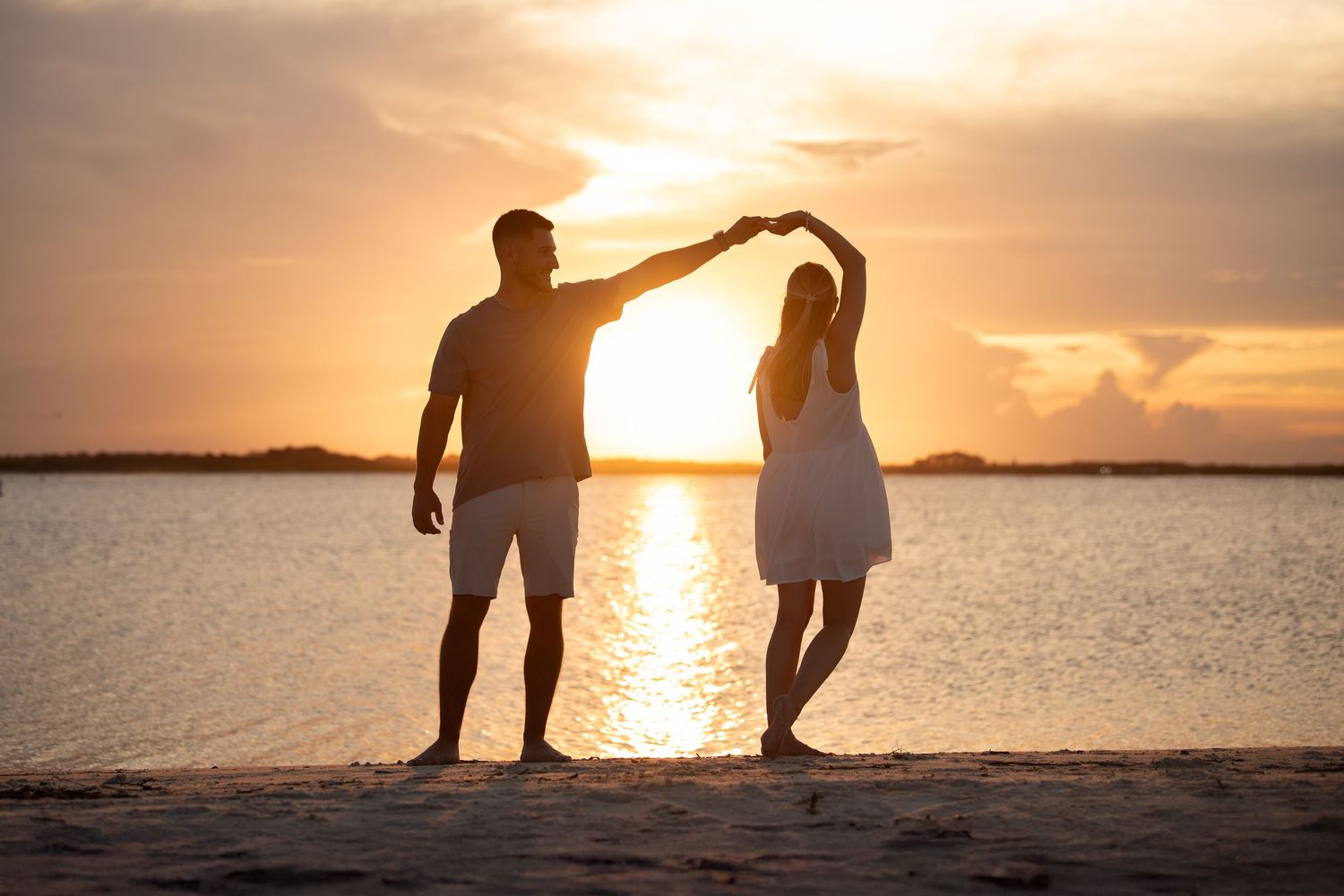 Romantic couple silhouetted against a golden sunset on a beach, holding hands raised in a heart shape.