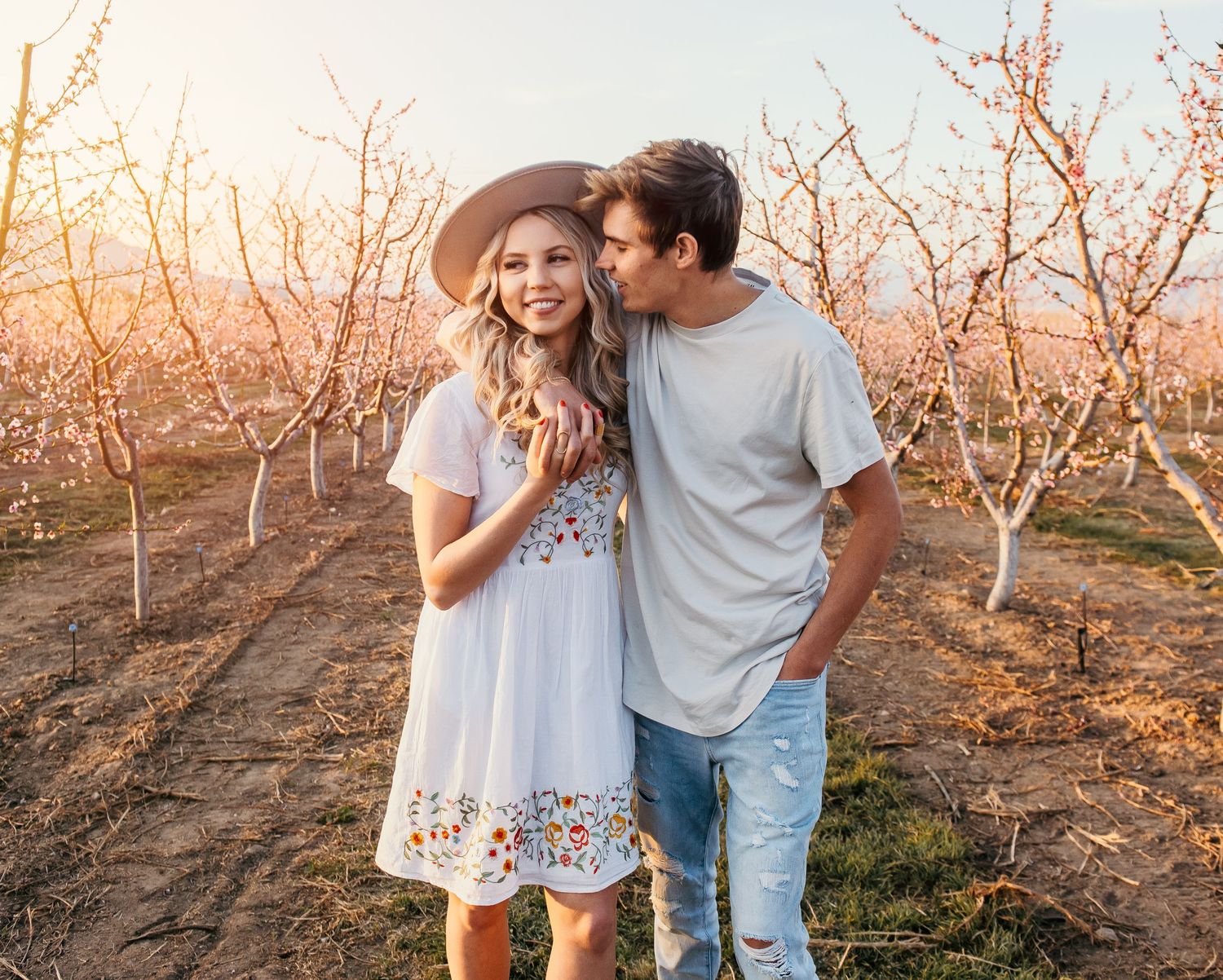 Couple embracing in blooming orchard at golden hour, woman in white floral dress and hat, man in gray tee and jeans.