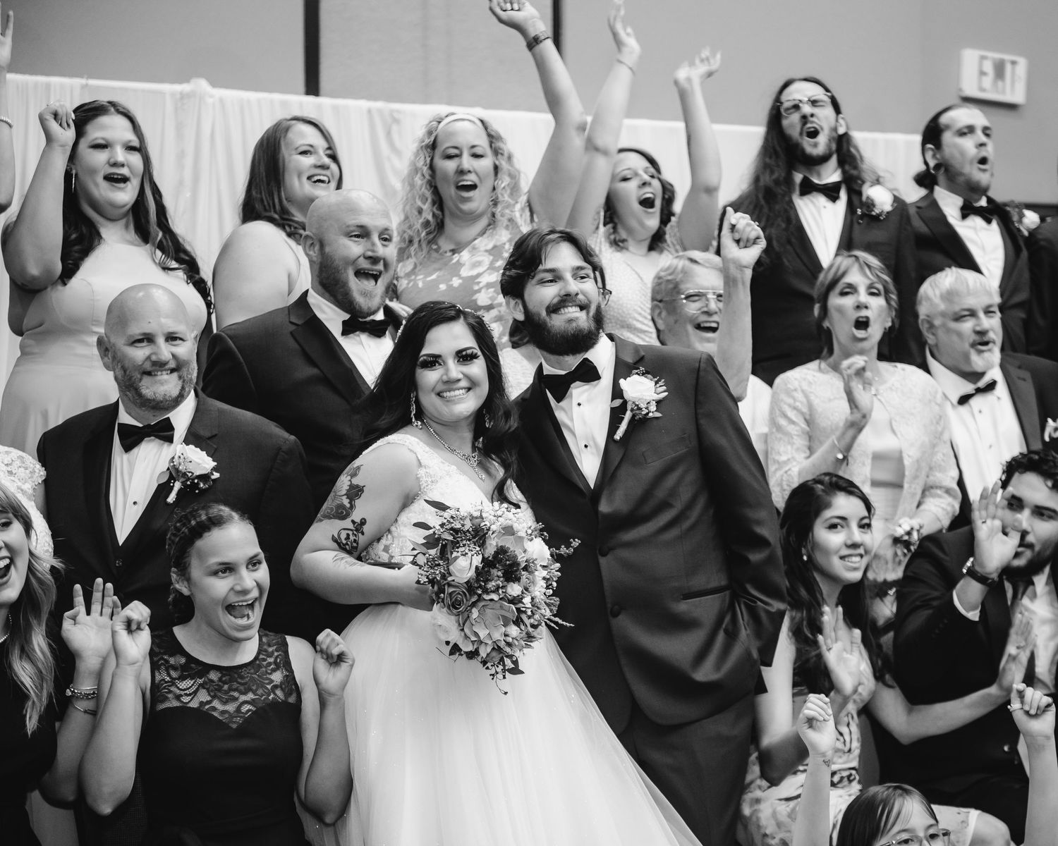 Black and white wedding photo of smiling bride and groom surrounded by cheering guests in formal attire.