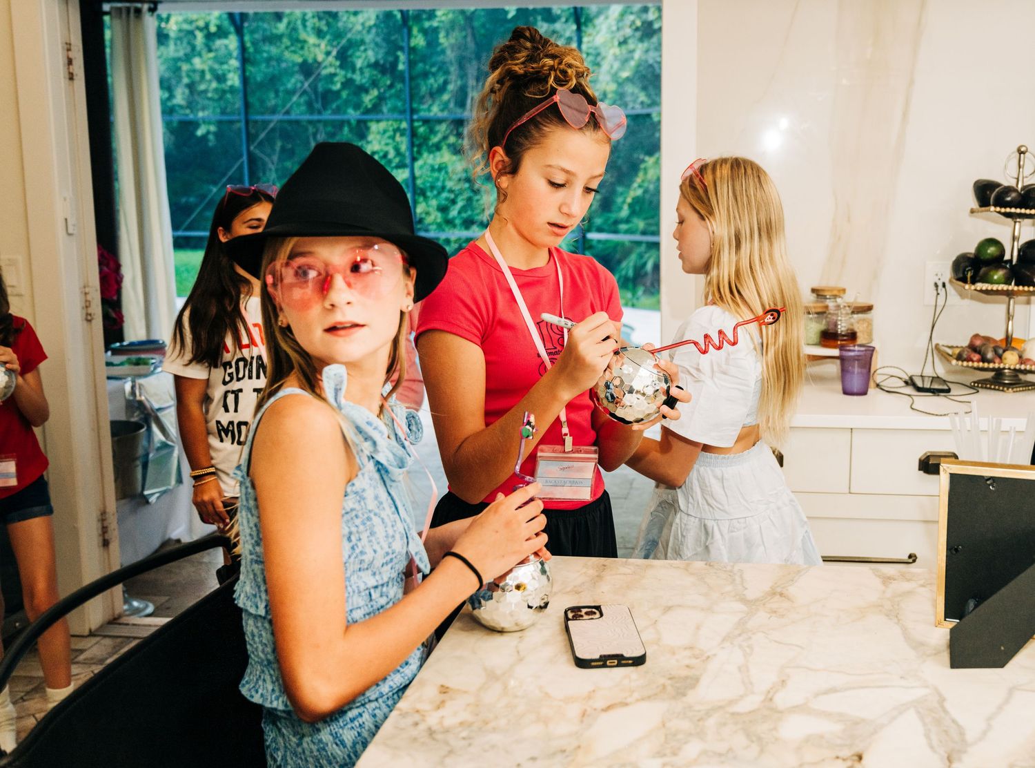 Girls gathered around a marble kitchen counter, one wearing a black hat and pink sunglasses, during a fun indoor activity.