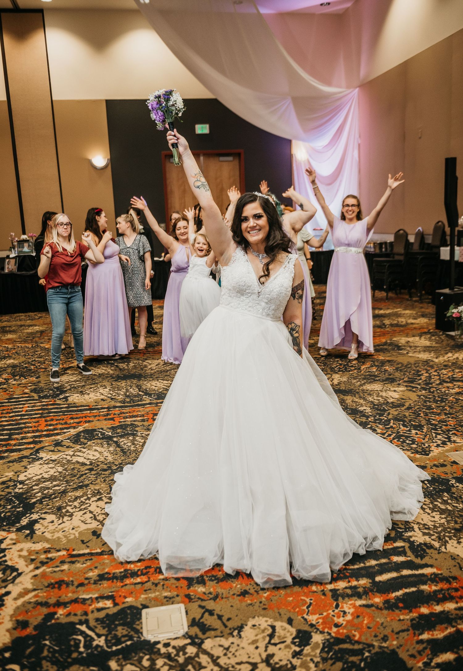Bride in white ballgown tosses bouquet at wedding reception with bridesmaids in lavender dresses cheering behind her.
