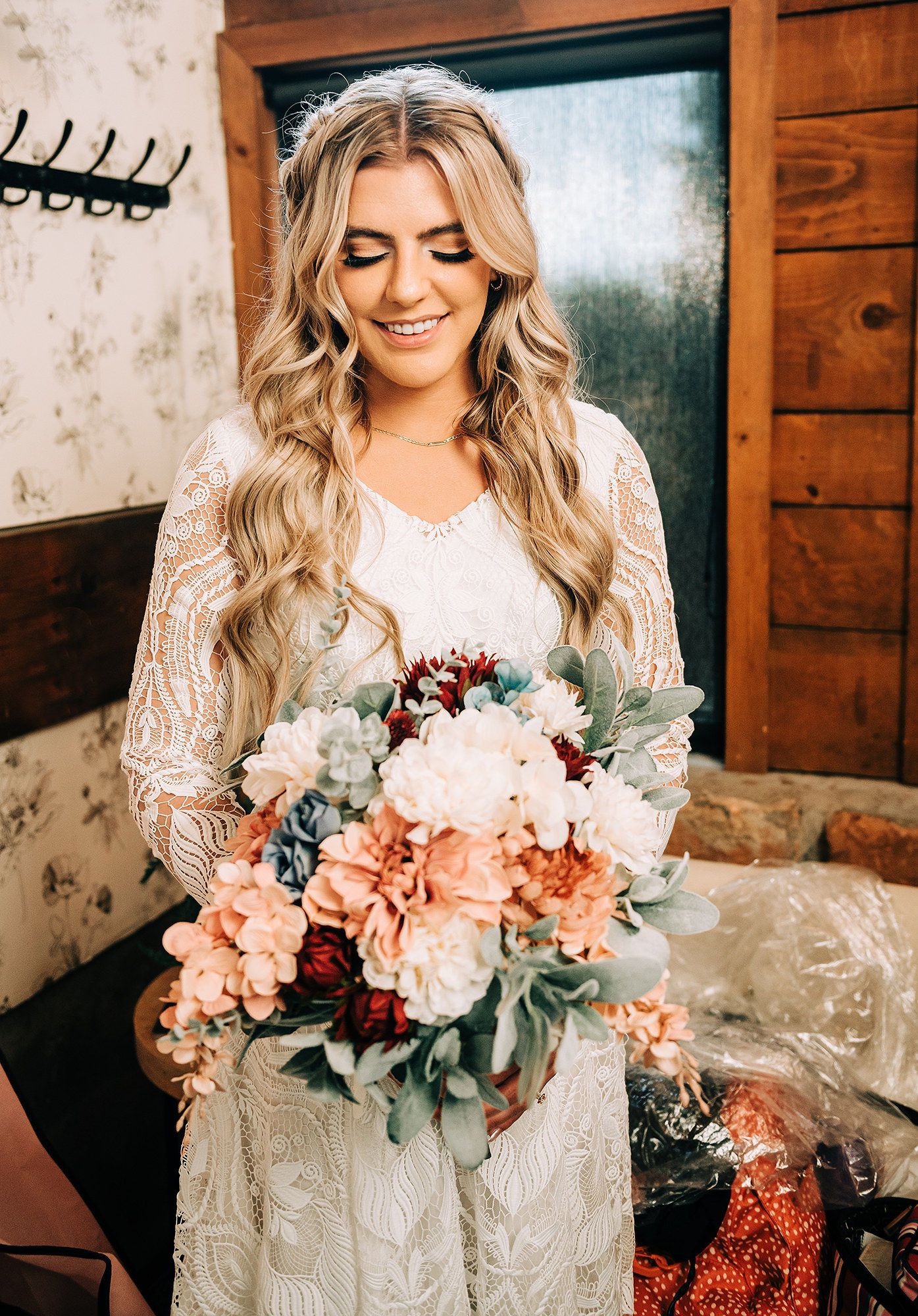 Smiling bride in lace long-sleeve gown holding a boho bouquet of peach, white, and burgundy flowers with eucalyptus.