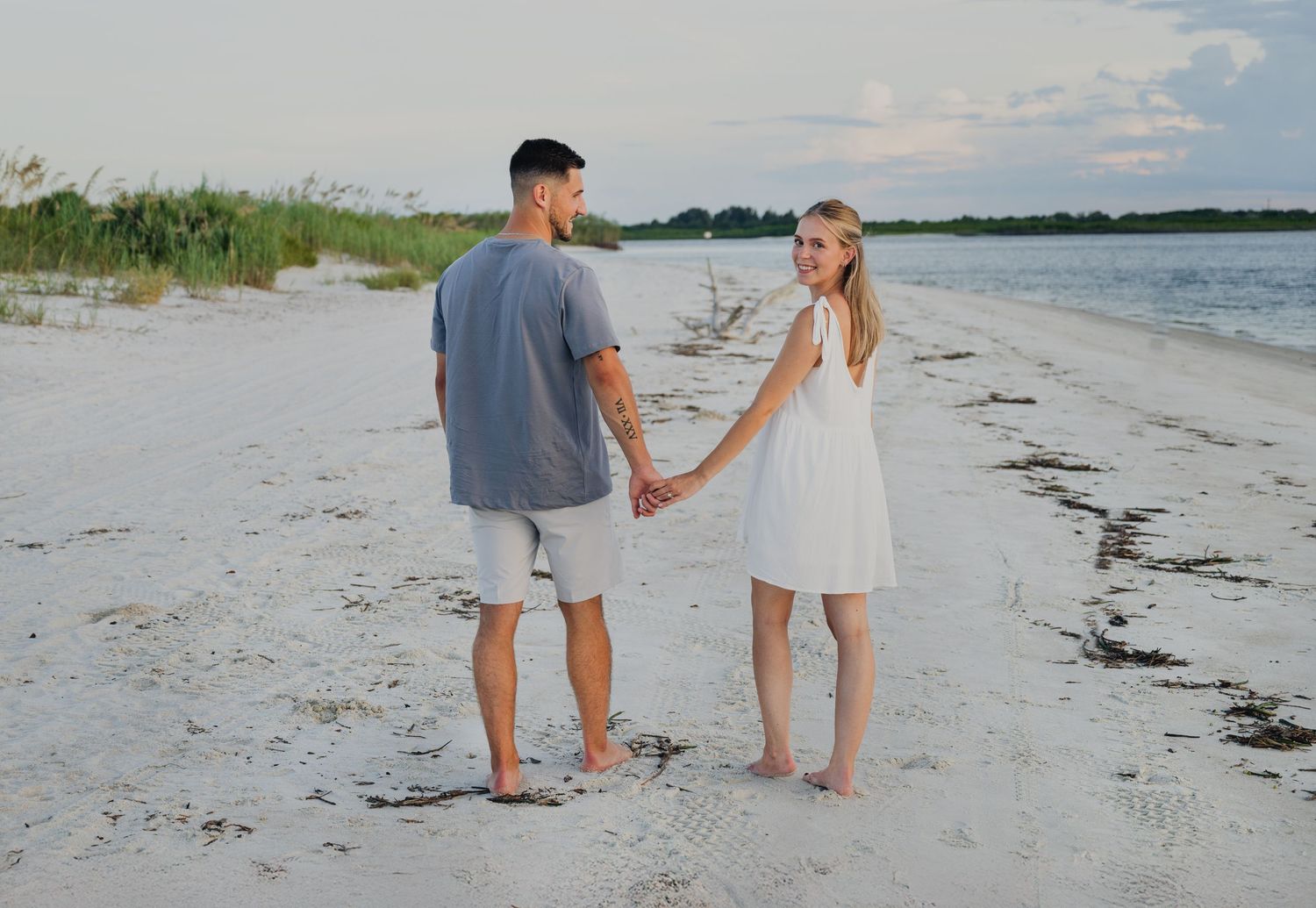 A couple holds hands on a white sandy beach, the woman in a white dress and man in gray, with calm water behind them.