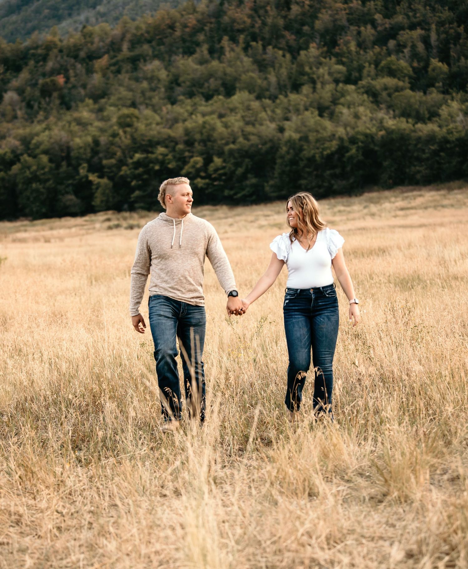Couple holding hands walking through golden field with forested mountain backdrop.