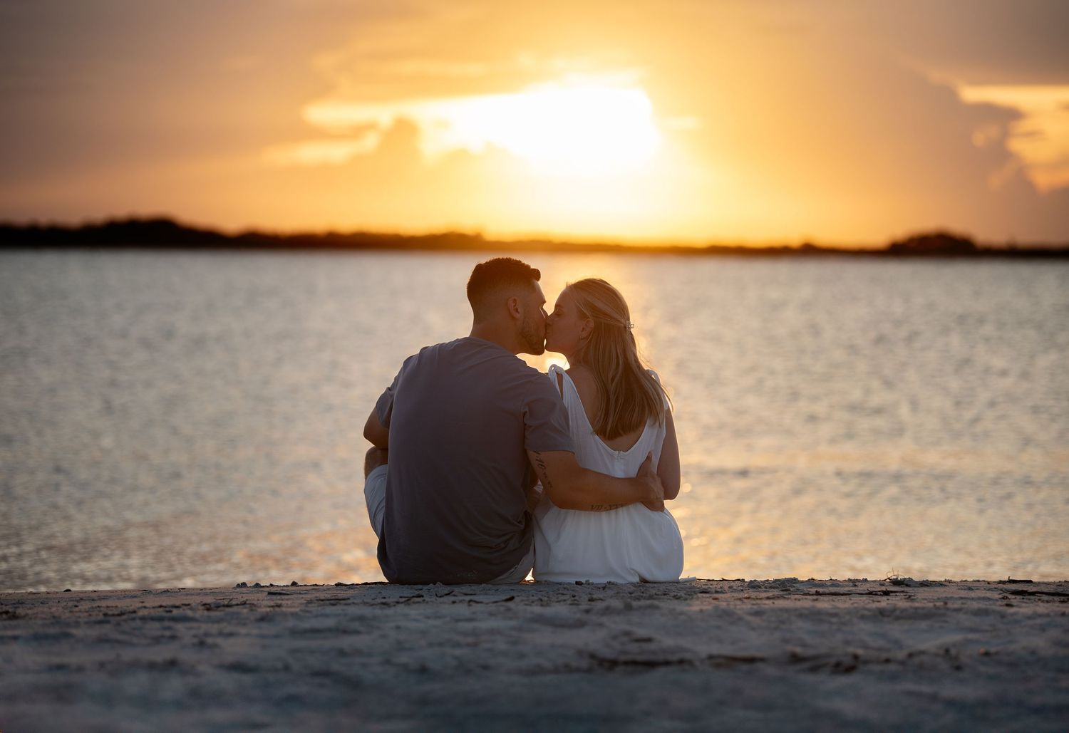 Couple sitting on beach shore kissing at golden sunset with calm water reflecting warm orange light.