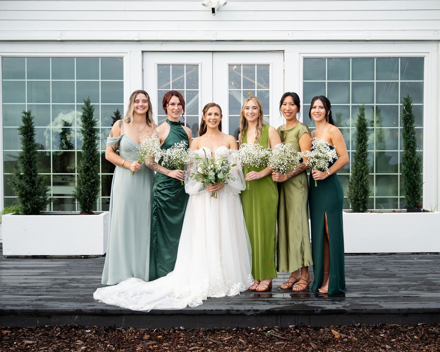 Bride in white gown poses with five bridesmaids in mixed green dresses, holding bouquets outside a white venue.