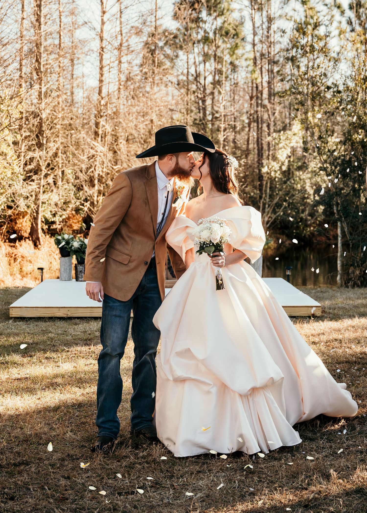 A couple shares a romantic kiss at an outdoor rustic wedding, groom in cowboy hat and bride in blush ball gown.