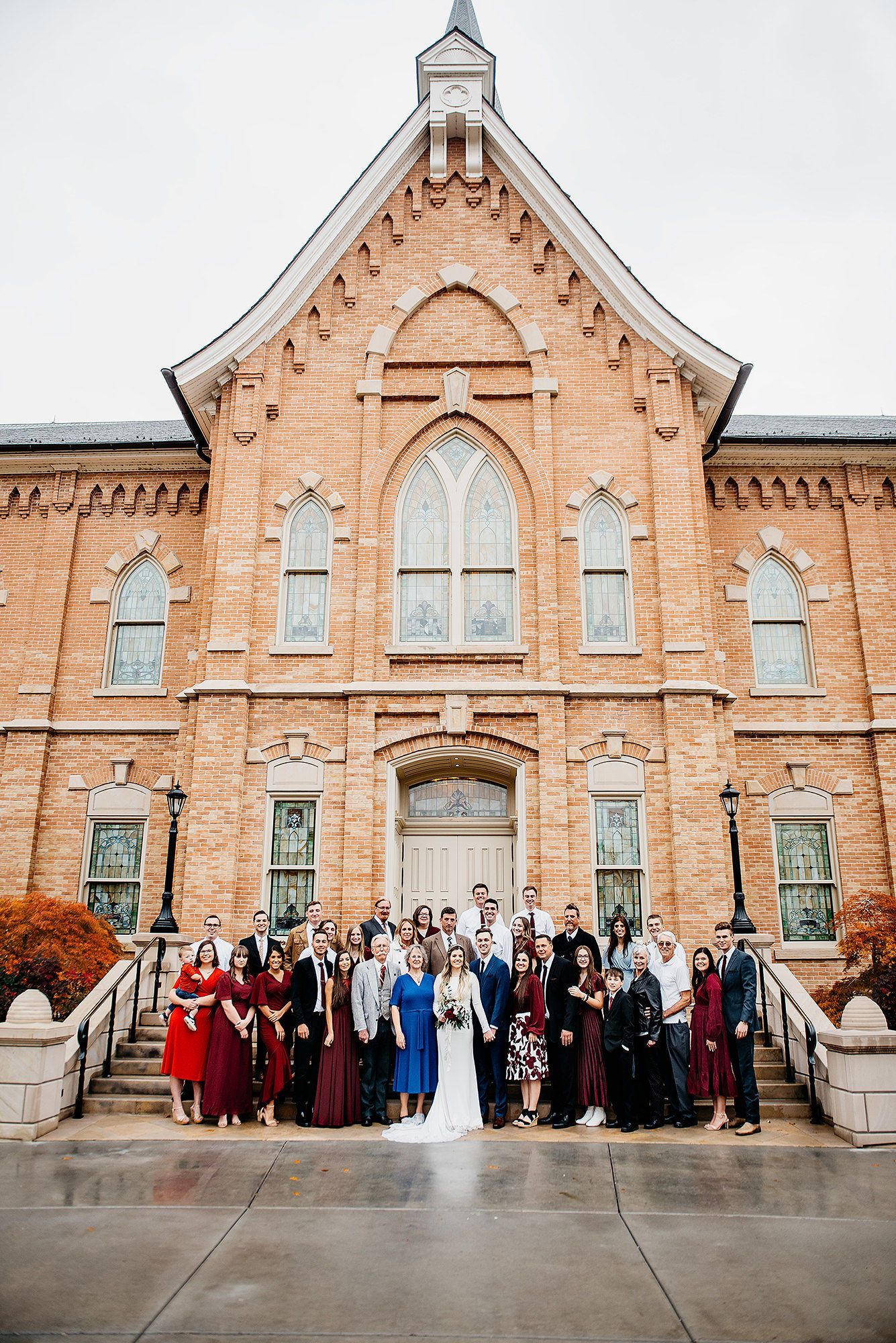 Wedding party poses in front of a historic Gothic brick church, bride in white gown surrounded by guests in burgundy.