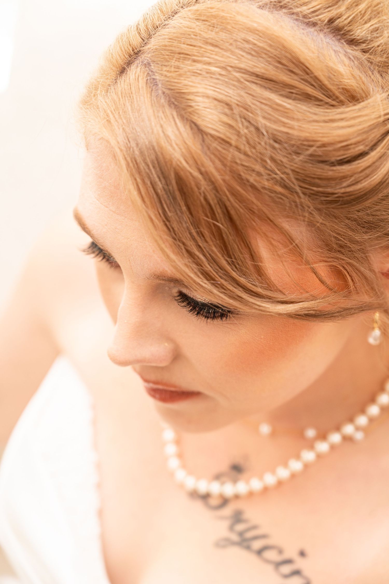 Close-up portrait of a blonde bride wearing pearl necklace and earrings with a tattoo on her chest, looking down.