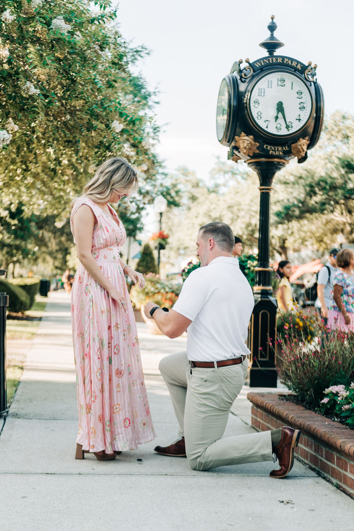 Man proposing on one knee to woman in pink floral dress near Winter Park Central Park clock tower.