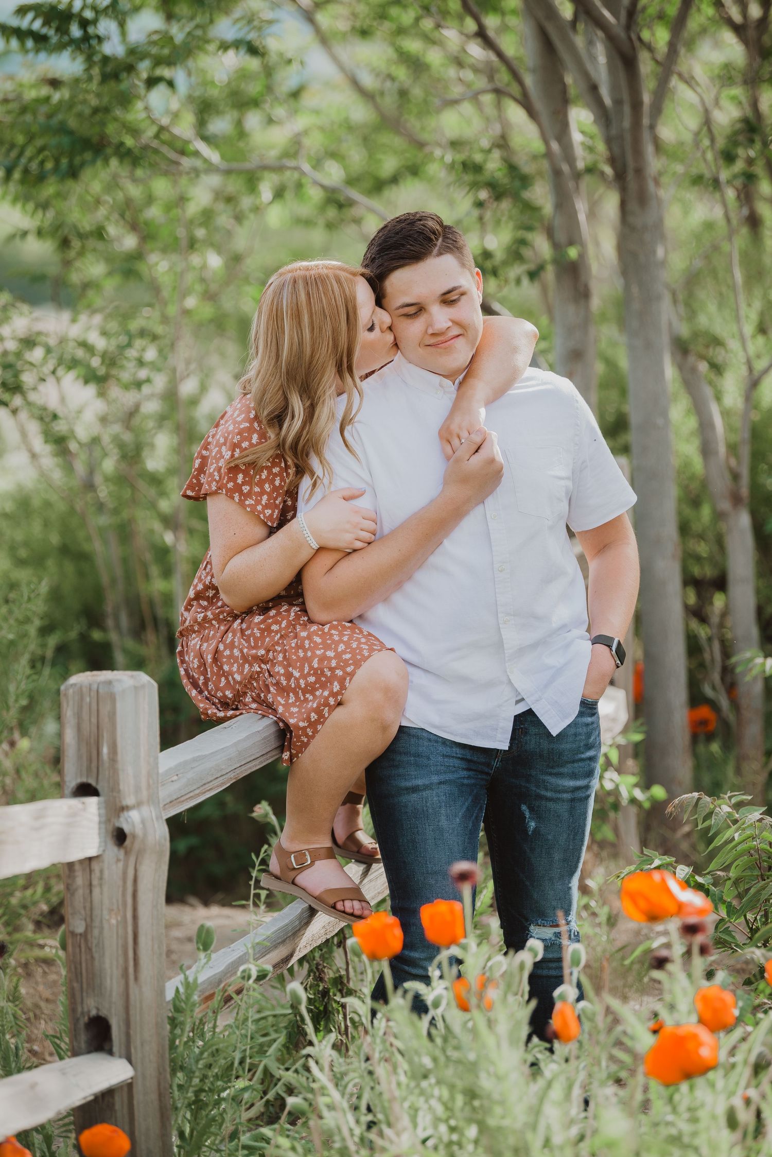 A couple shares a romantic moment outdoors, woman kissing mans cheek near a wooden fence with orange poppies blooming nearby.