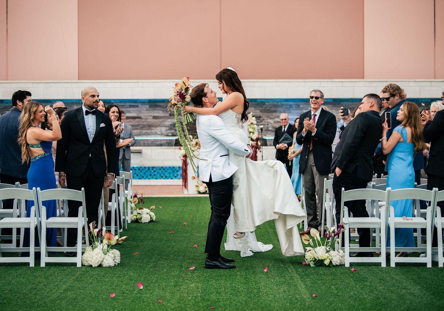 Bride and groom share first kiss at outdoor wedding ceremony with guests applauding in background.