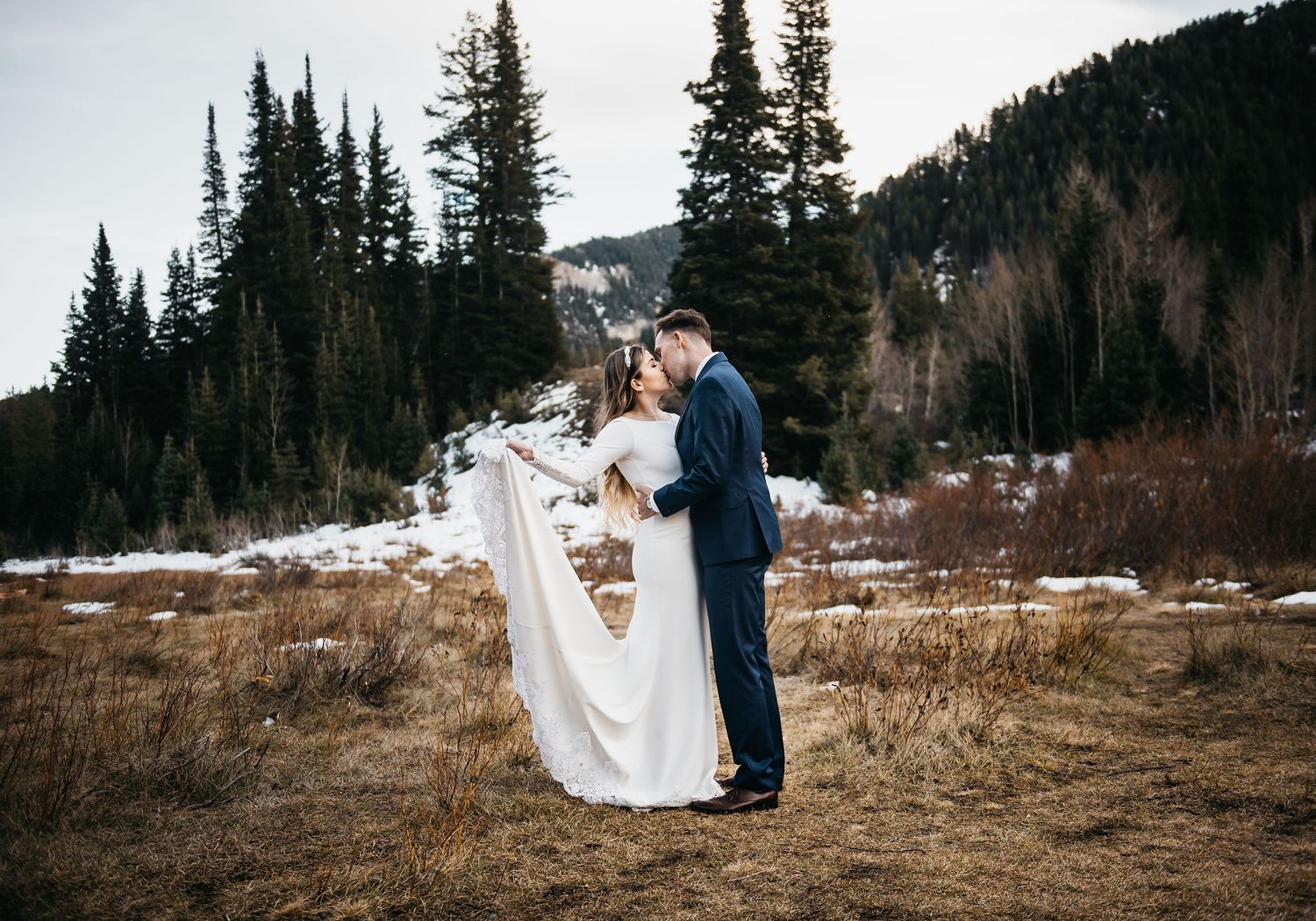 Couple sharing a romantic kiss in snowy mountain meadow, bride in white gown, groom in navy suit, evergreen trees backdrop.