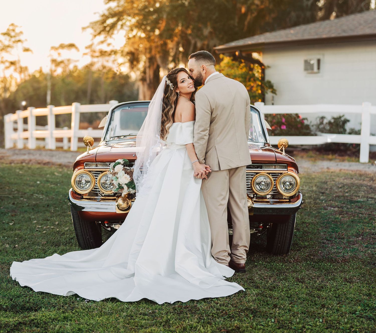 Bride and groom posing together in front of a vintage car at golden hour on their wedding day.