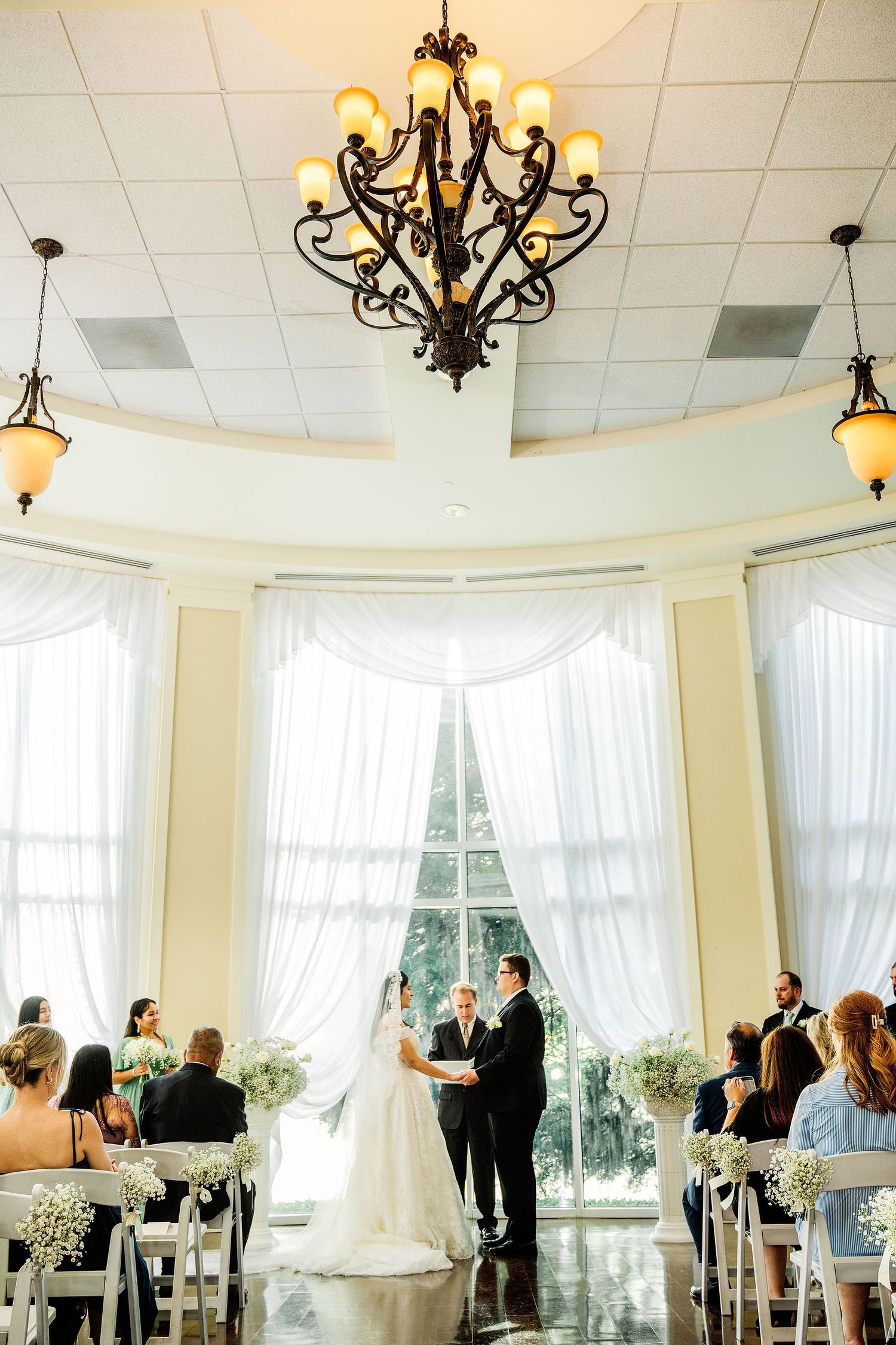 Elegant indoor wedding ceremony with couple at altar, white drapes, chandelier, and floral arrangements in a bright venue.