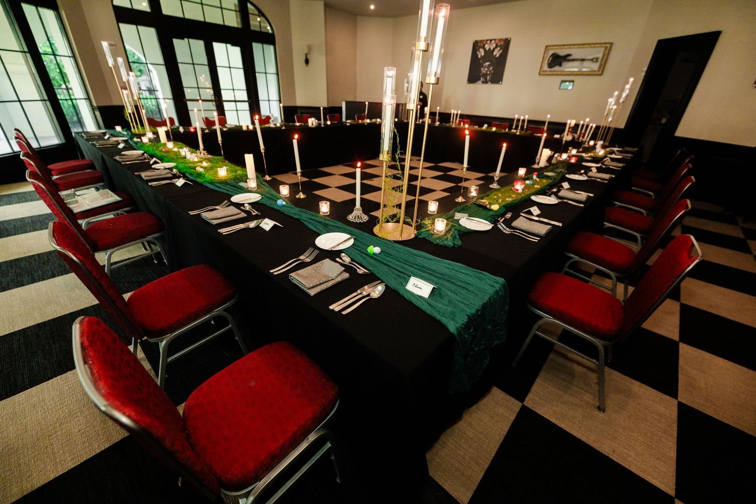 Elegant banquet hall with red chairs, black tablecloths, green runner, tall gold candelabras, and checkered floor.