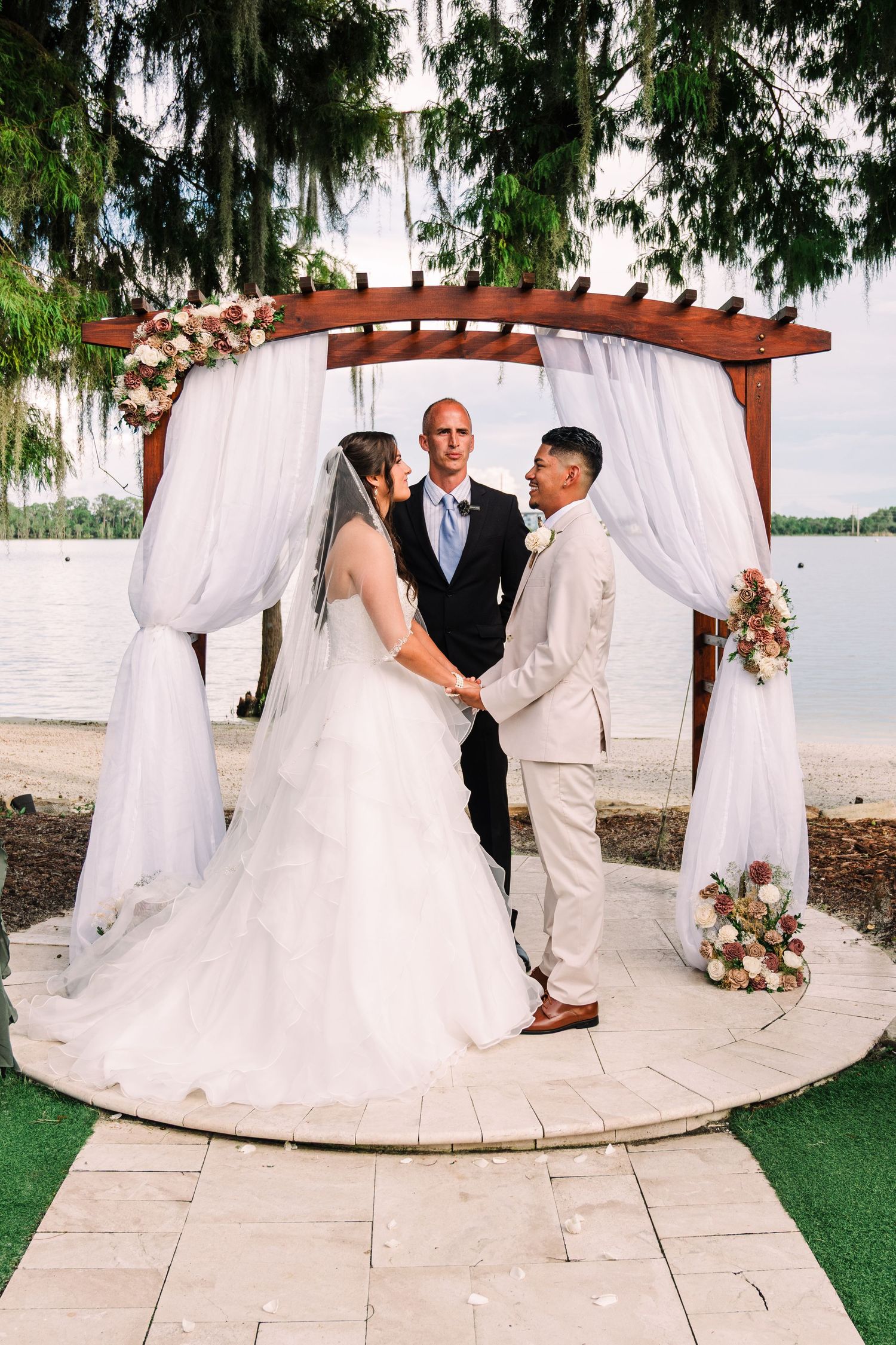 Couple exchanging vows at outdoor waterfront wedding ceremony under white draped pergola with floral arrangements.