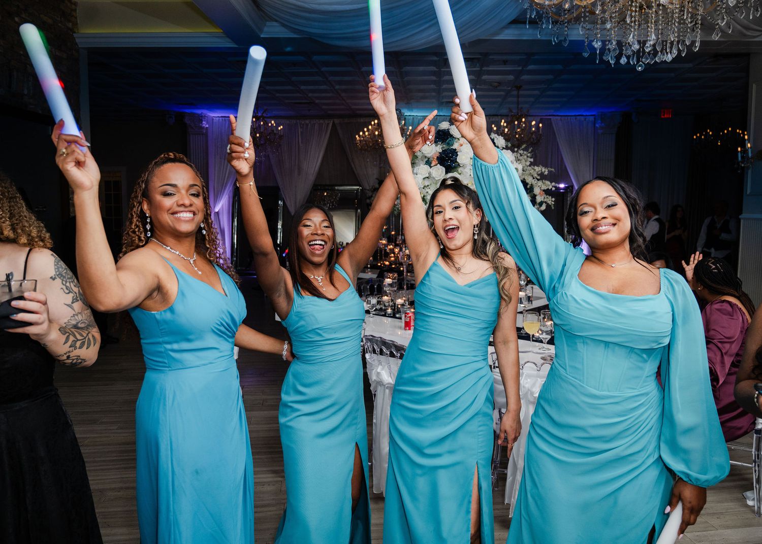 Four bridesmaids in matching teal gowns celebrate at a wedding reception, raising colorful glow sticks in the air.