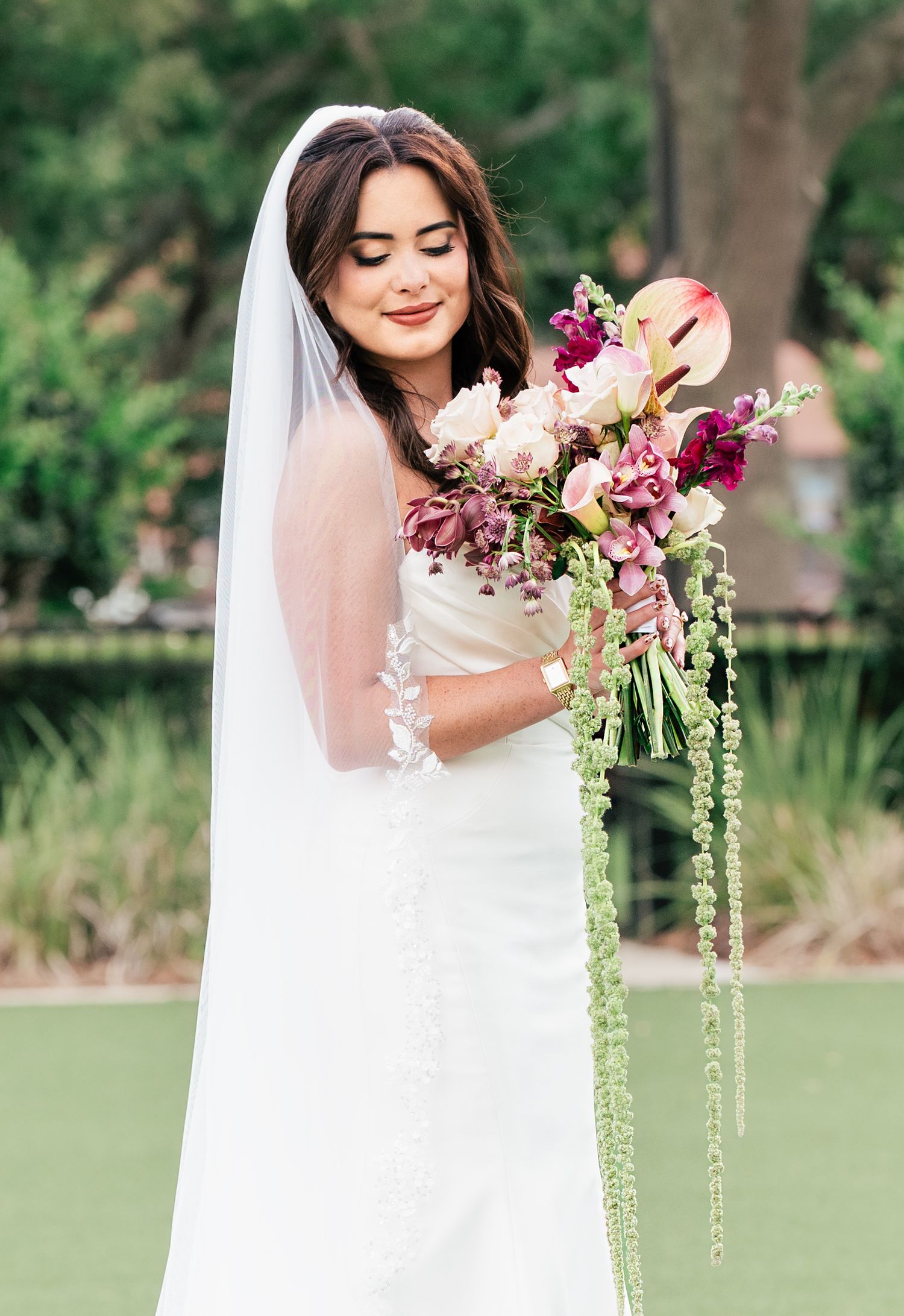 Bride in white gown and veil holding a cascading bouquet of pink, burgundy, and green flowers outdoors.