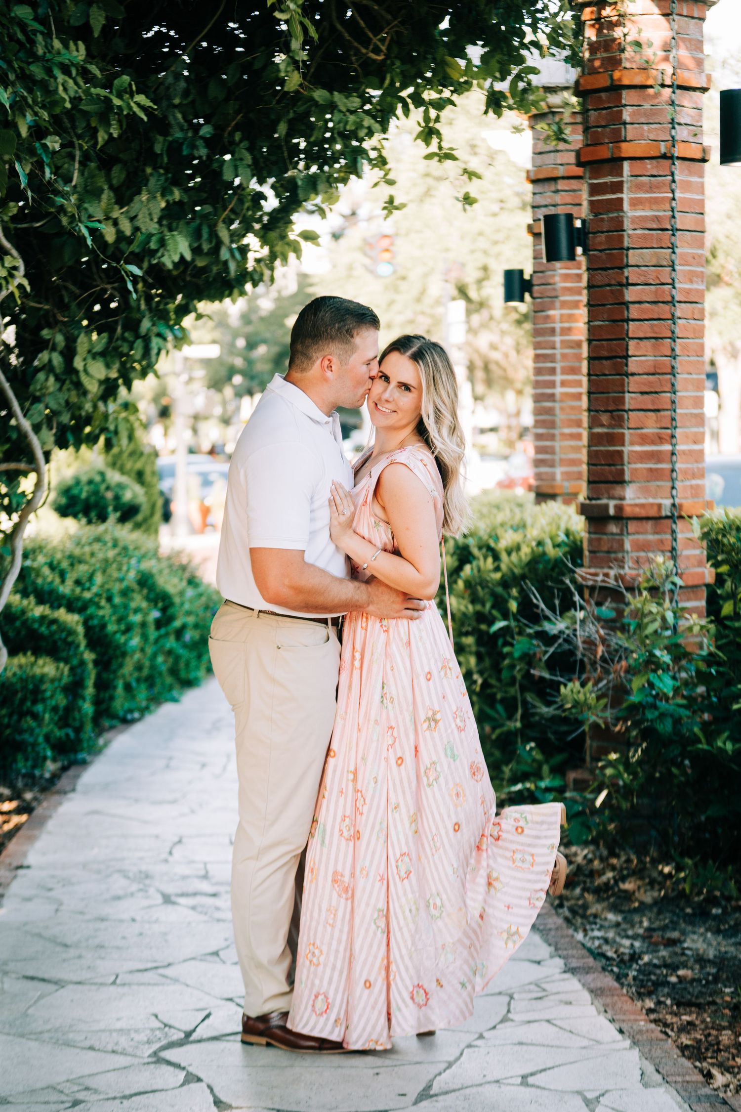Couple embracing on a stone path surrounded by lush greenery, woman in a flowing pink dress, man in white shirt.