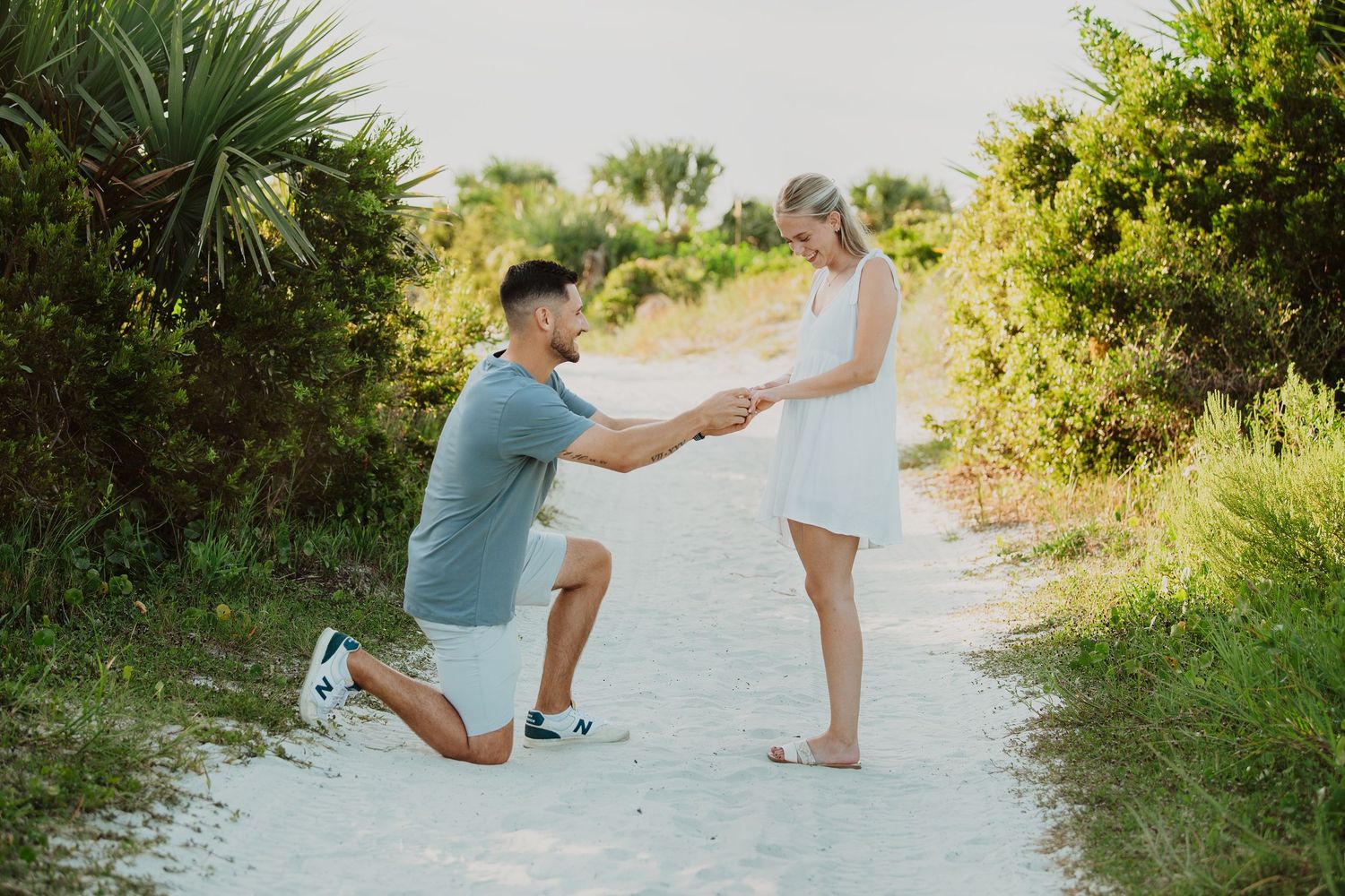 Man kneeling on sandy path proposing to woman in white dress surrounded by lush green tropical foliage.